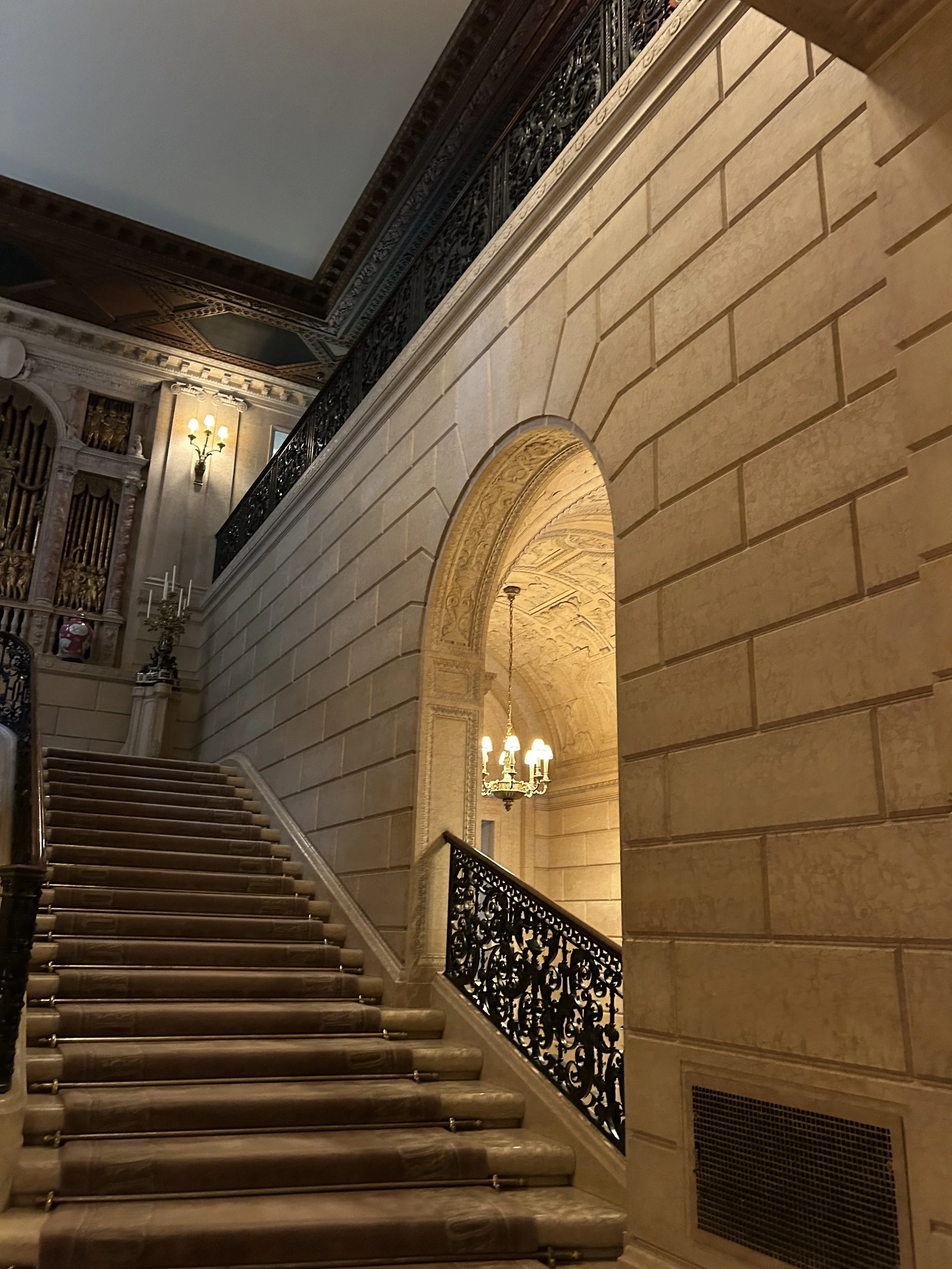 Elegant staircase with marble steps, ornate black wrought-iron railings, and classic wall-mounted chandeliers in a luxurious interior.