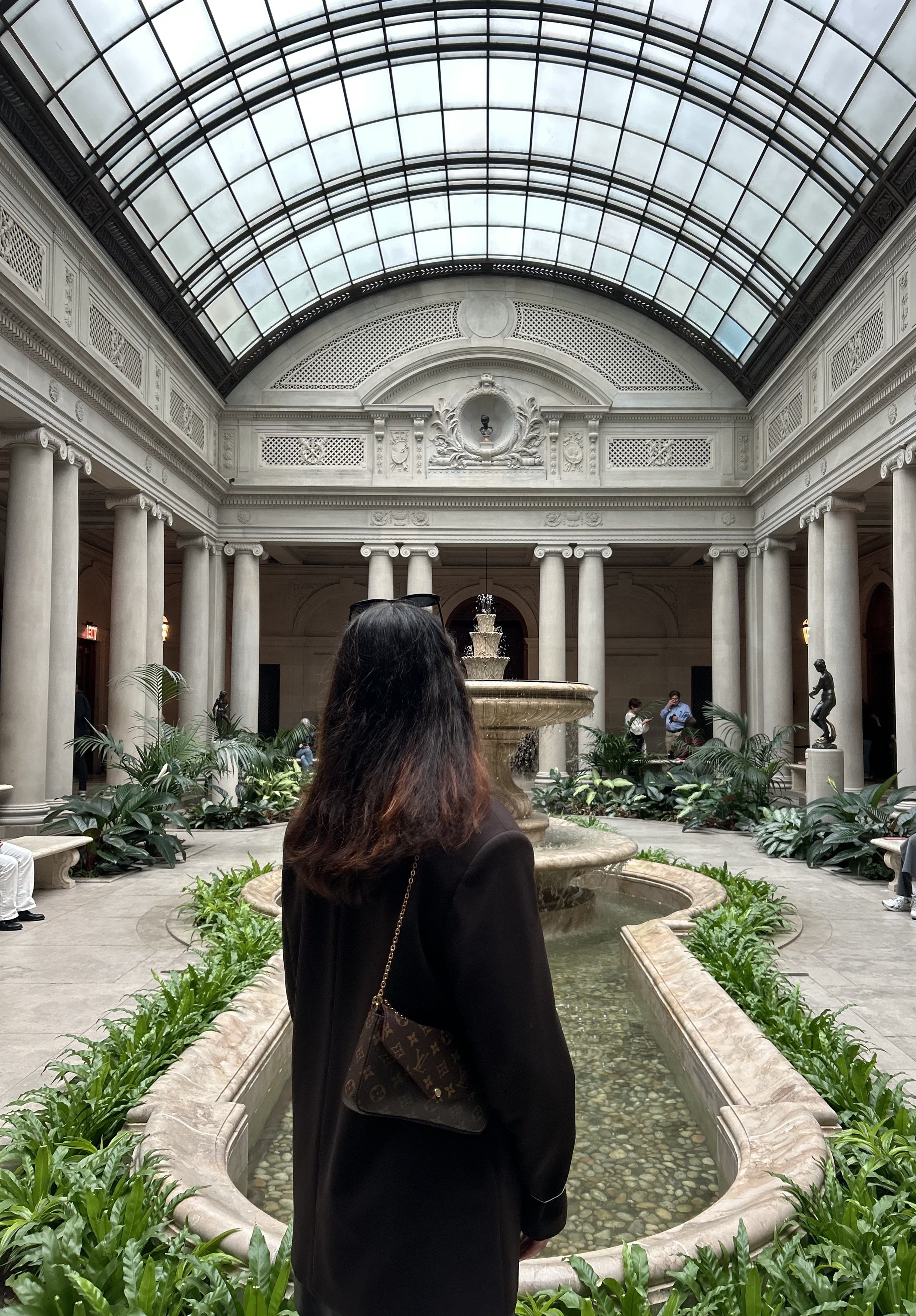 Interior of a grand shopping arcade or historic building with marble columns, a glass dome ceiling, and a central fountain surrounded by greenery. Several people are in the background, and a woman with dark hair and a handbag stands in the foreground