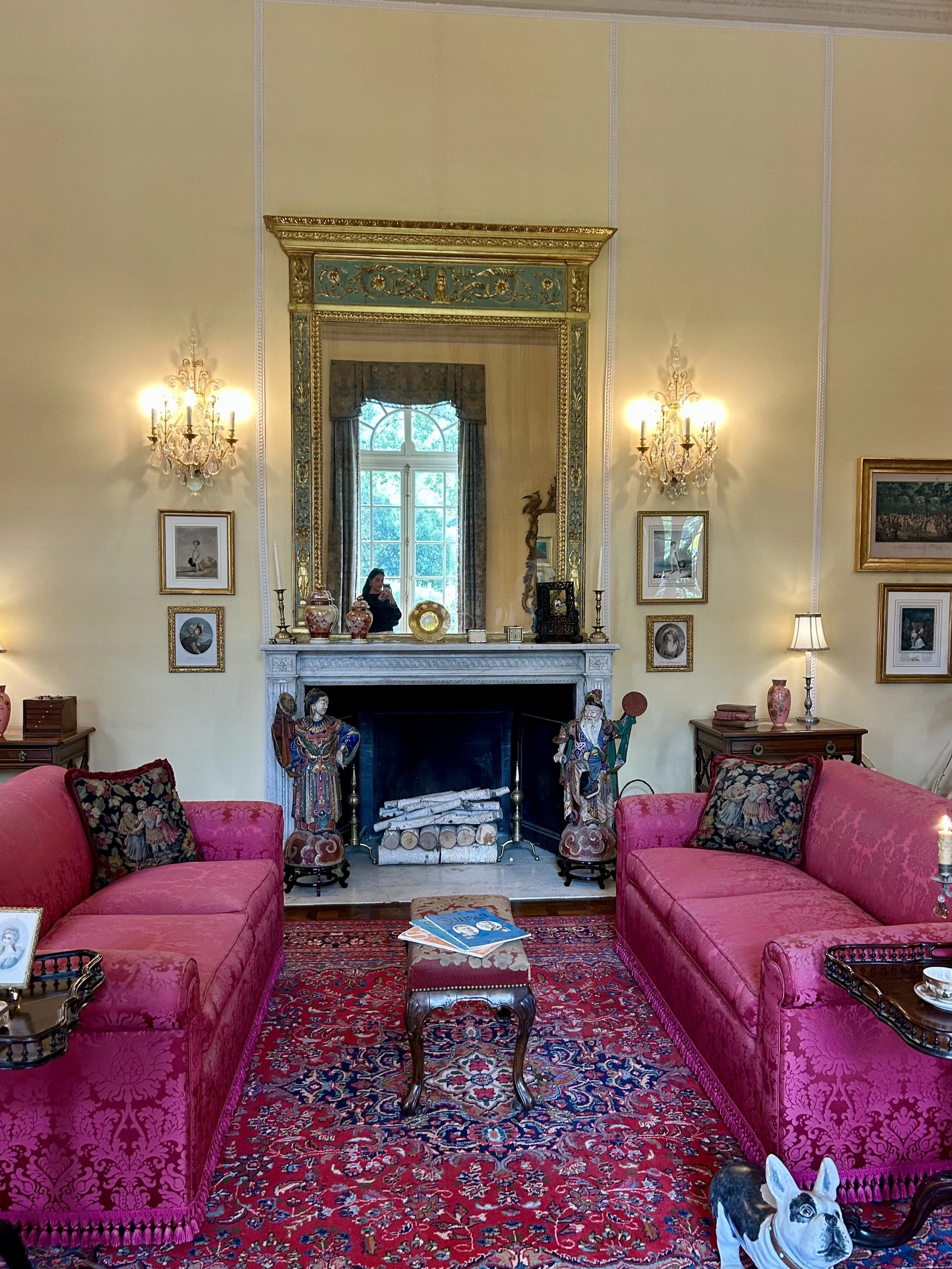 A cozy living room with pink velvet sofas, a patterned Persian rug, and a white marble fireplace. The mantle holds decorative vases and figurines. An ornate gold-framed mirror hangs above the fireplace, reflecting a large window with drapes. Two wall