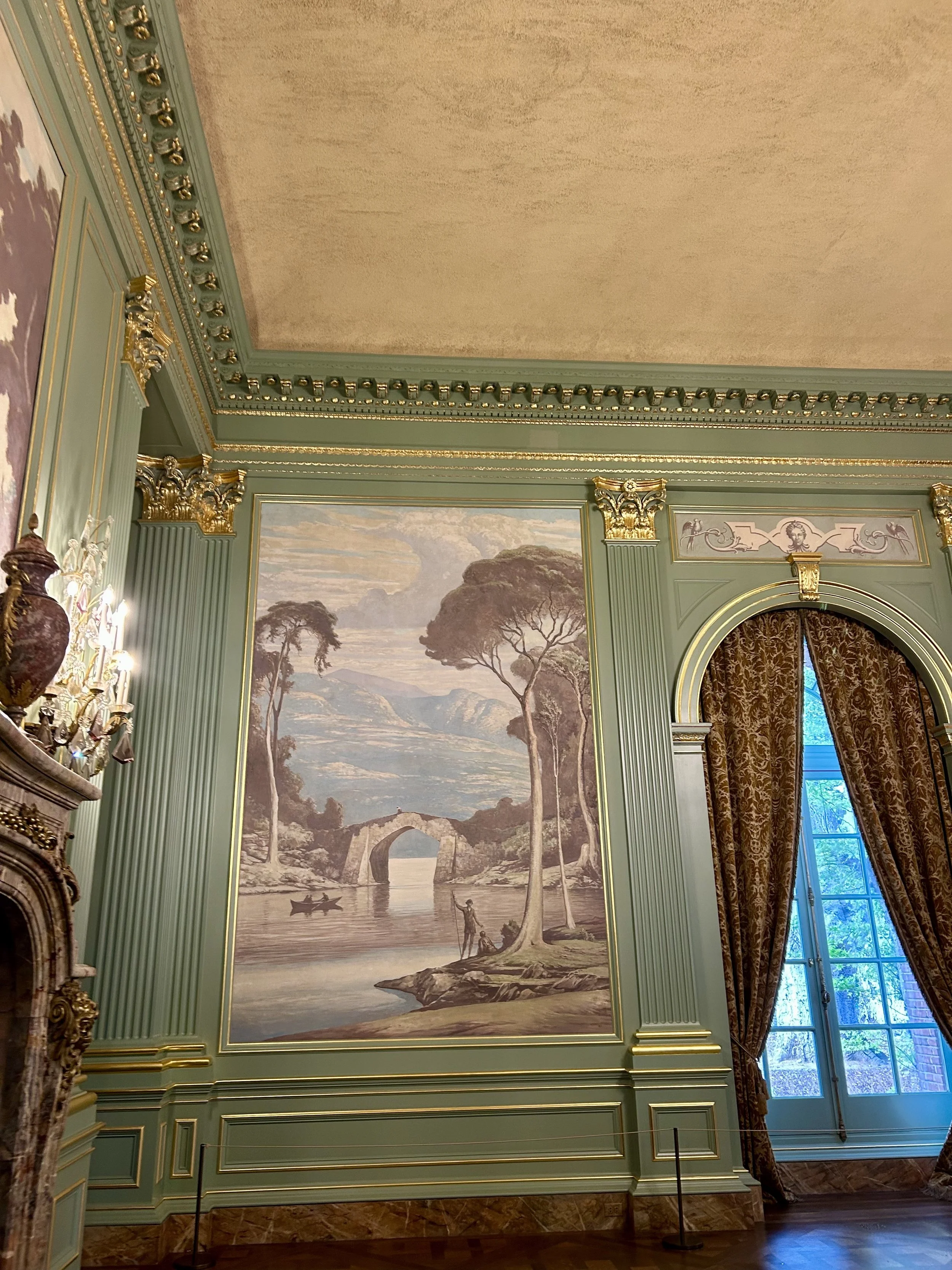 An ornate room with light green walls decorated with gold accents, featuring a large landscape mural of trees, water, and mountains, with a window draped with patterned curtains on the right side.