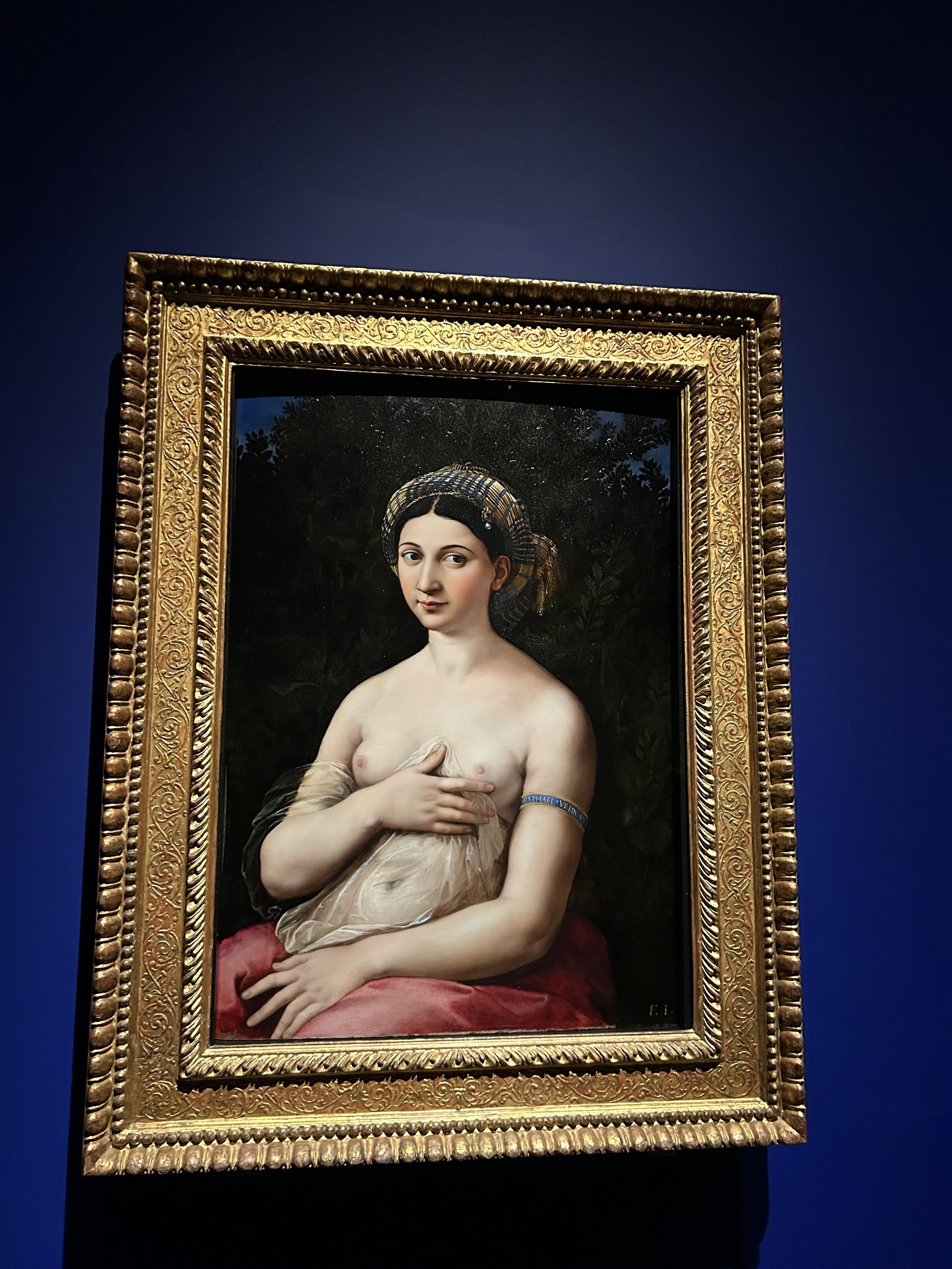 Painting of a woman with dark hair, wearing a striped head covering, holding her bare chest with one hand, framed in an ornate gold frame, displayed against a dark background.