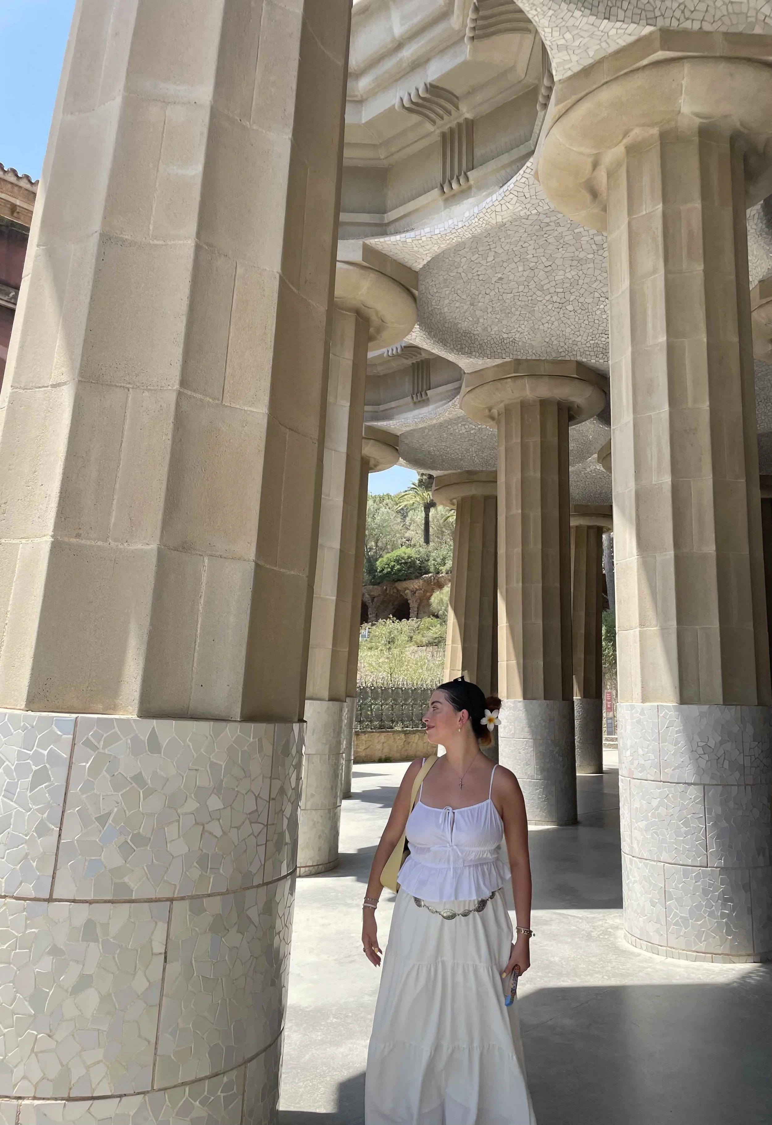A woman in a white dress with a flower in her hair, standing under a structure with stone pillars and curved, textured ceilings, outdoors with trees in the background.