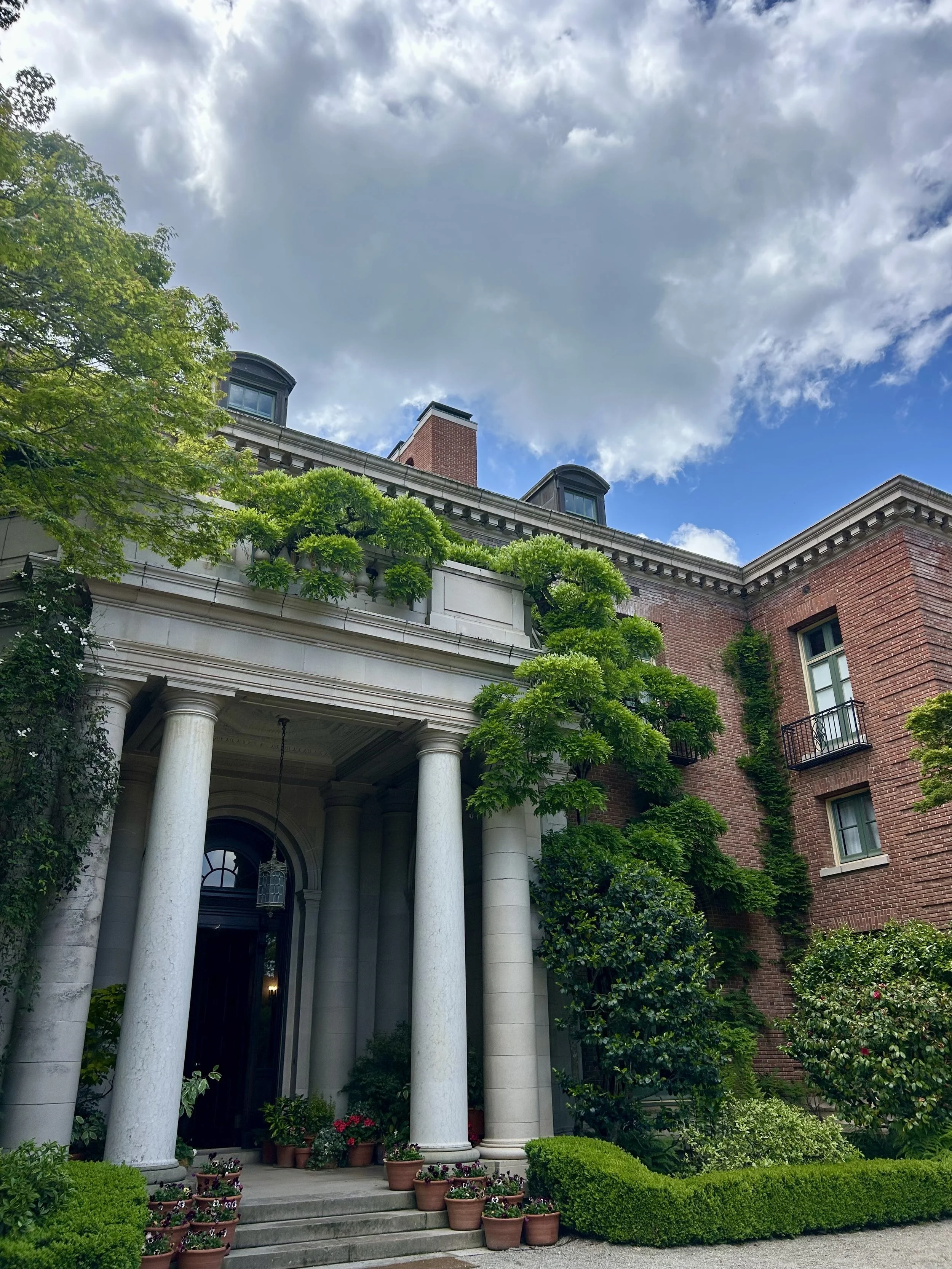 A historic brick building with elegant white columns at the entrance, surrounded by lush green trees, potted flowers, and a well-maintained garden under a partly cloudy sky.