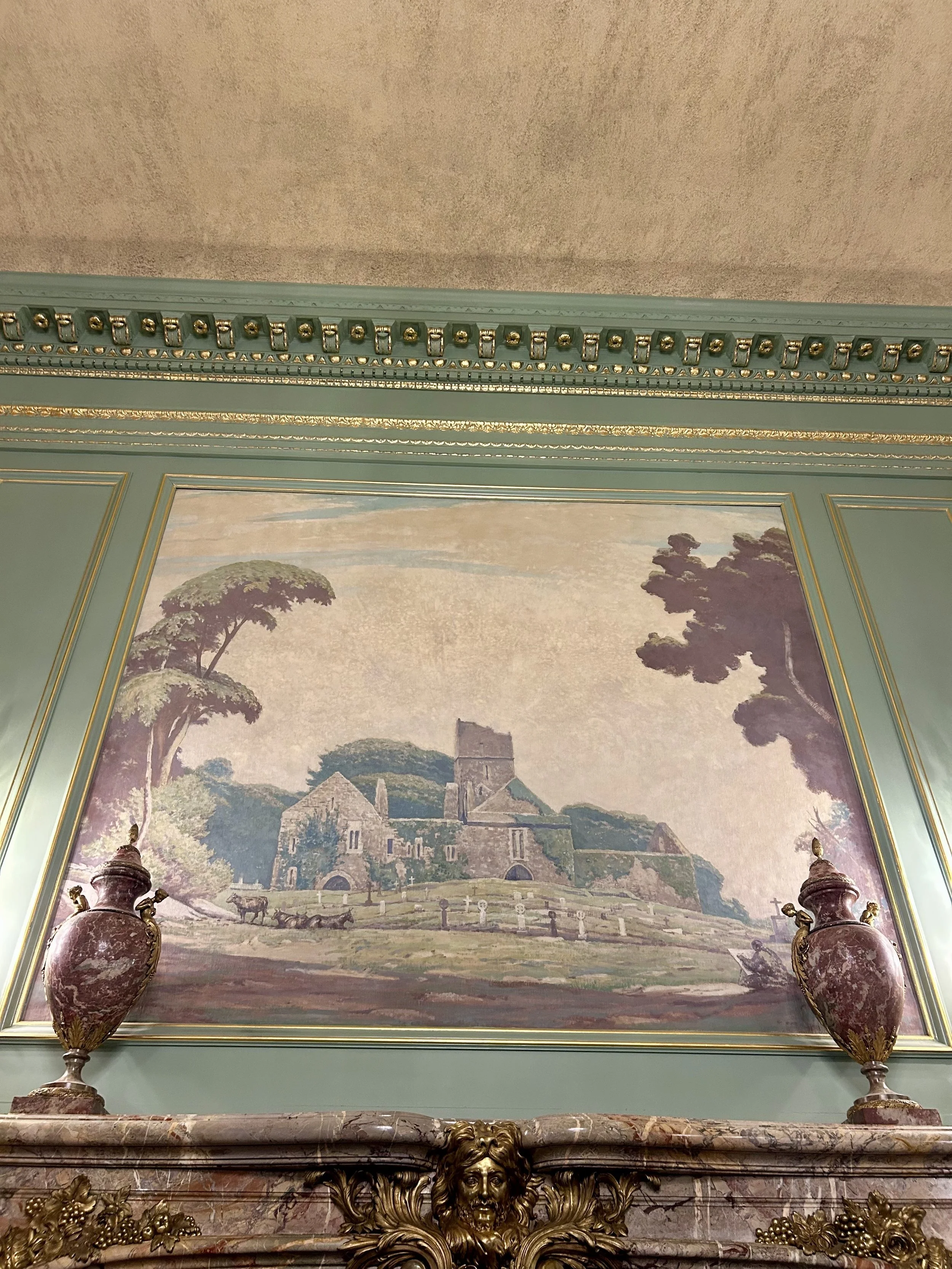 Decorative wall with a landscape painting of a castle and trees, framed in green with gold accents. Below the painting is a marble mantelpiece featuring a sculpted lion's head and ornate gold floral decorations. On each side of the mantel stand tall,