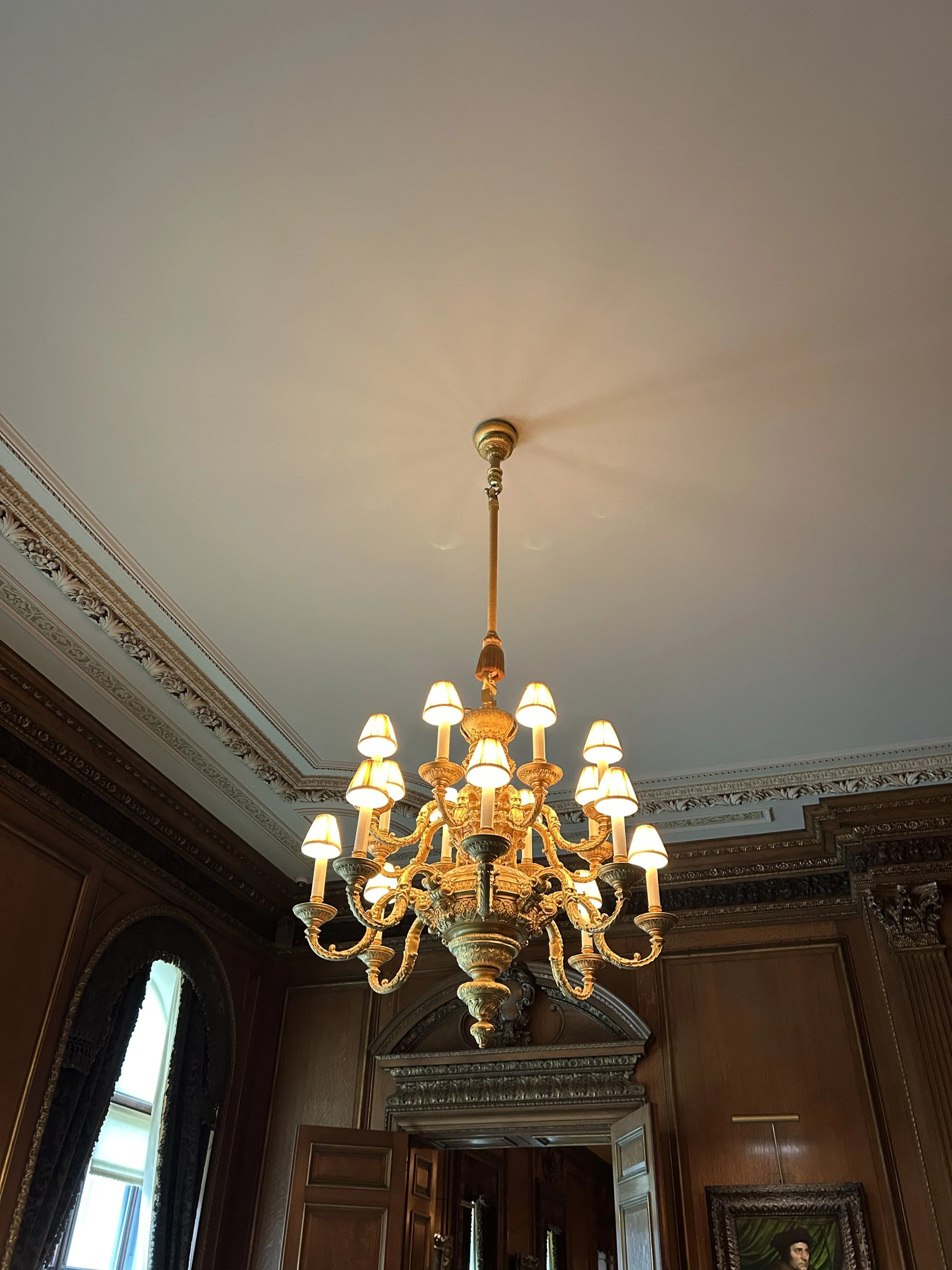 A grand chandelier with multiple lights hangs from a ceiling in a richly decorated room. The ceiling features ornate crown molding, and the walls are wood-paneled with arched windows and framed artwork.
