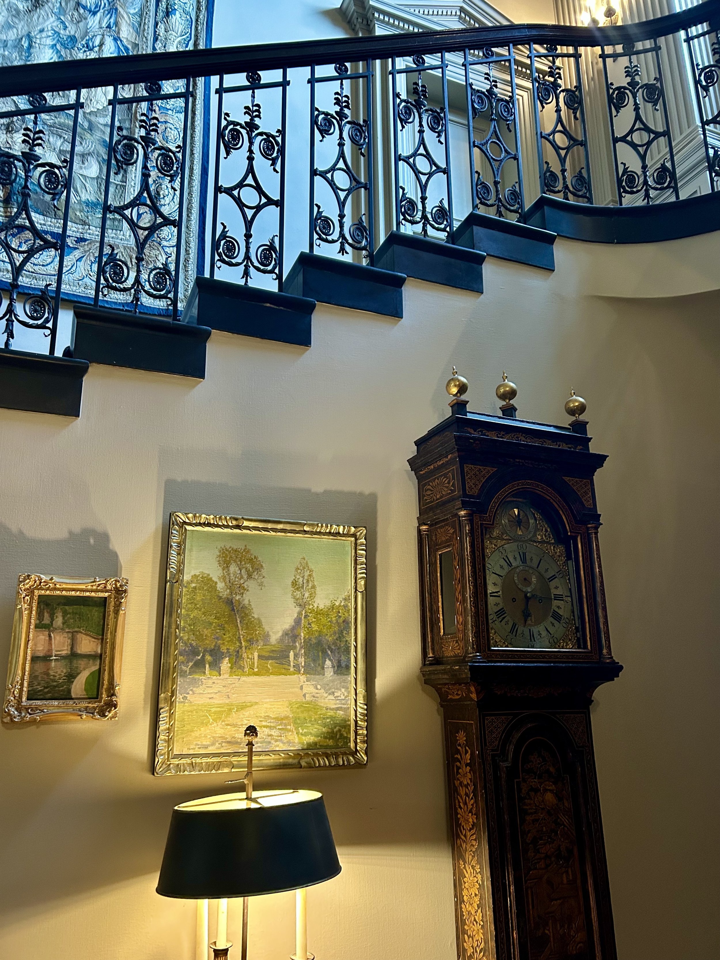 Interior view of a room with an ornate vintage grandfather clock, framed paintings on the wall, a decorative railing on the upper level, and a table lamp with a black lampshade in the foreground.