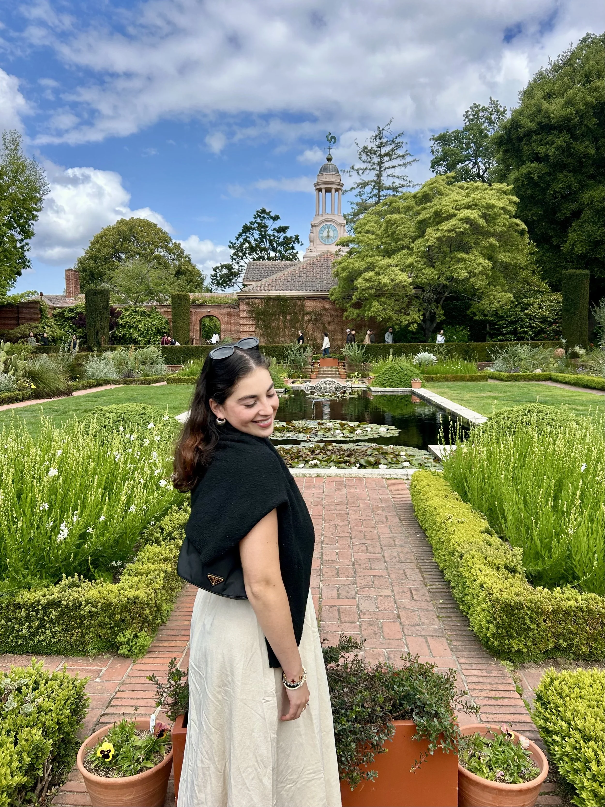 A smiling woman with dark hair and sunglasses on her head, dressed in a light-colored dress and black shawl, standing in a lush garden with a brick path, potted flowers, green bushes, trees, and a fountain, with a brick building and clock tower with 