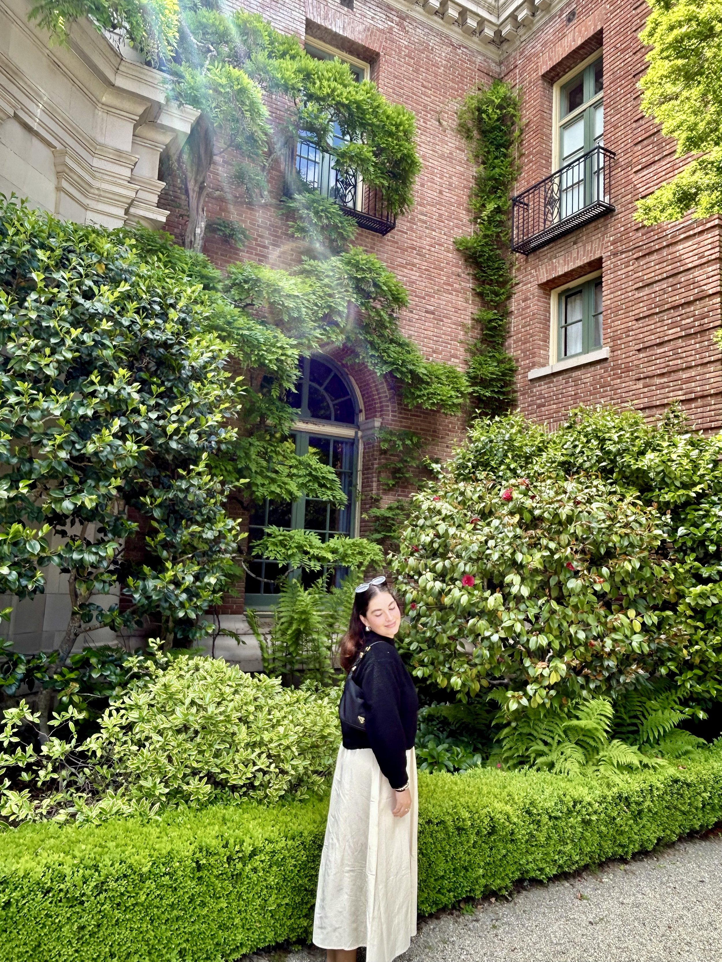 A woman with long dark hair, wearing a black jacket and white skirt, stands in front of lush green bushes and trees outside a brick building with decorative architectural details and small balconies with black railings.