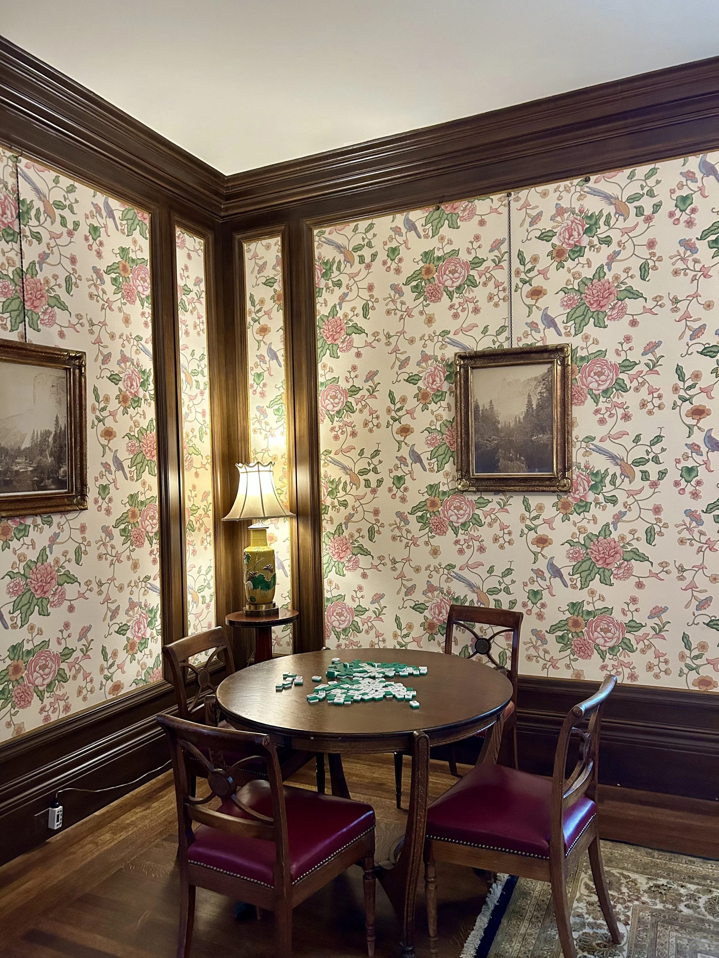 A round wooden table with scattered Mahjong tiles, surrounded by four matching chairs with burgundy cushions, in a room with floral wallpaper, wood trim, a lamp, and framed pictures on the wall.
