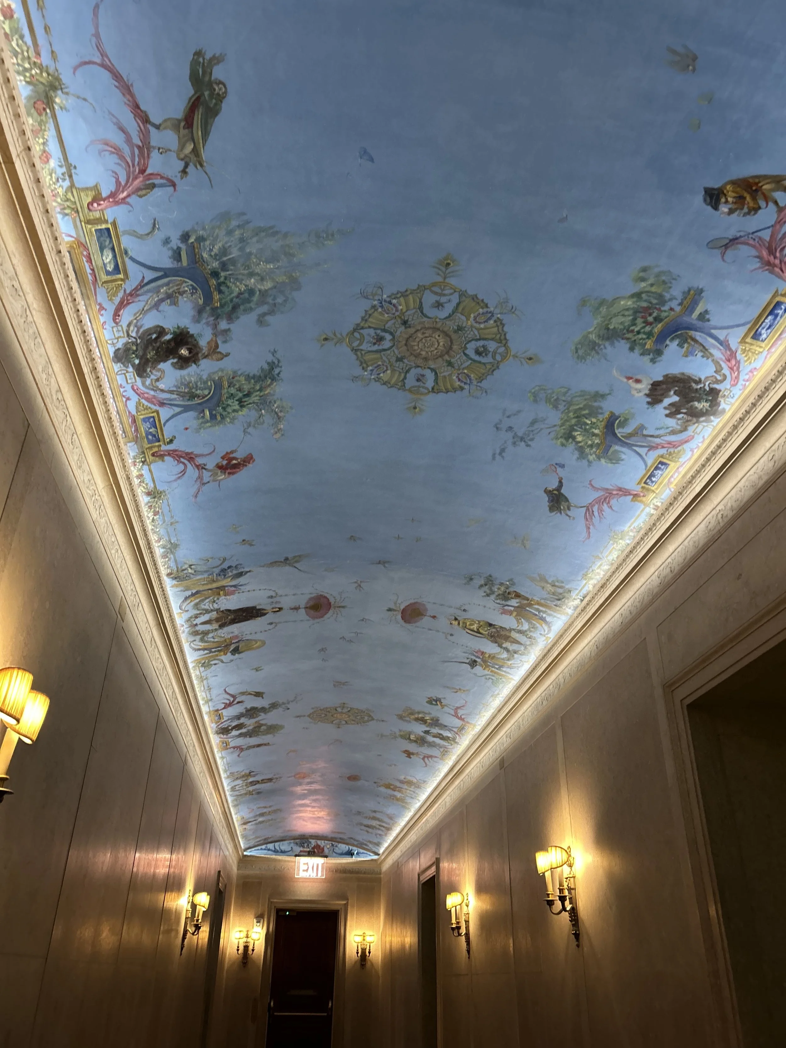 Decorative ceiling with a mural of figures, trees, and ornate designs, illuminated by wall lamps in a hallway with wood-paneled walls and an exit sign.