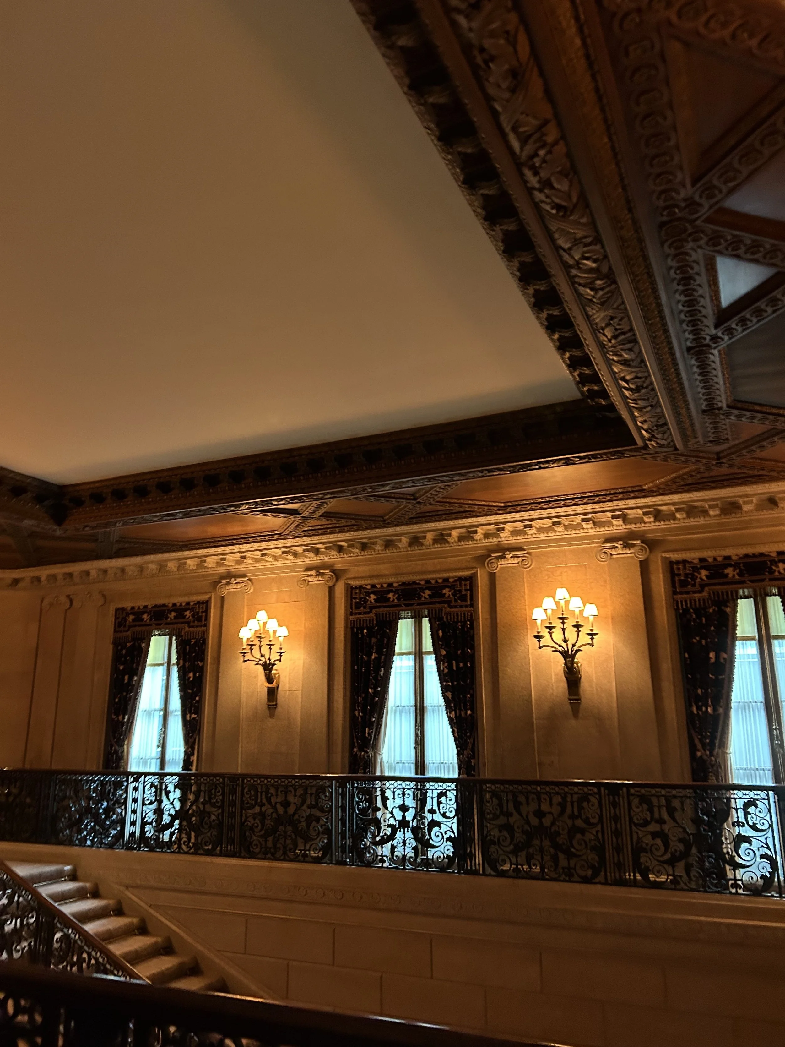 Elegant interior room with ornate crown molding and wall sconces, tall windows with curtains, and a decorative wrought iron railing along a staircase.