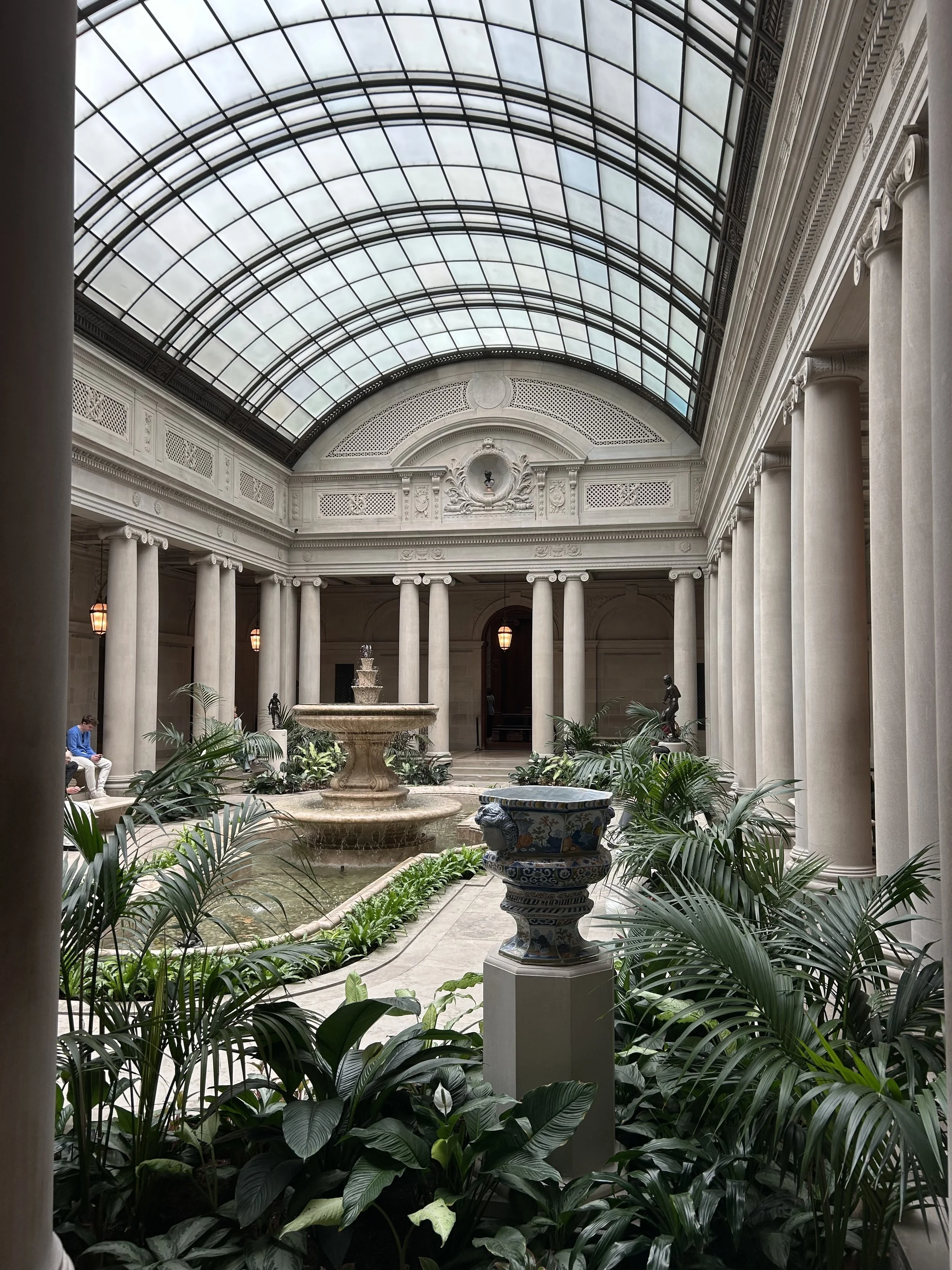 Indoor courtyard with a glass arched ceiling, classical columns, a fountain, and lush green plants.