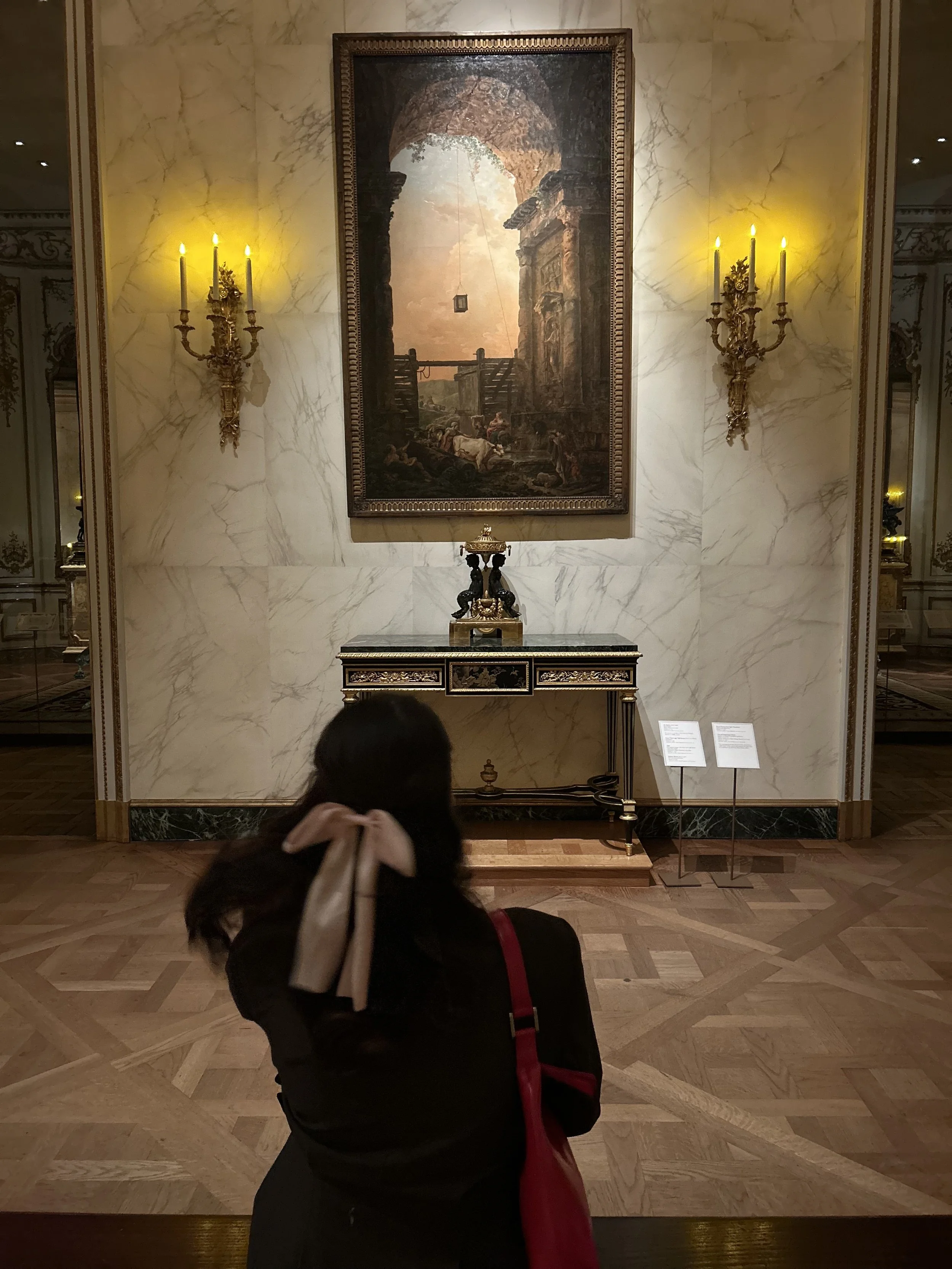 A museum interior featuring a large framed painting of an ancient ruin with a sunset sky, flanked by ornate wall sconces with lit candles, and a small decorative table with a golden sculpture below the painting. A person with dark hair tied back and a red bag is in the foreground, partially visible.