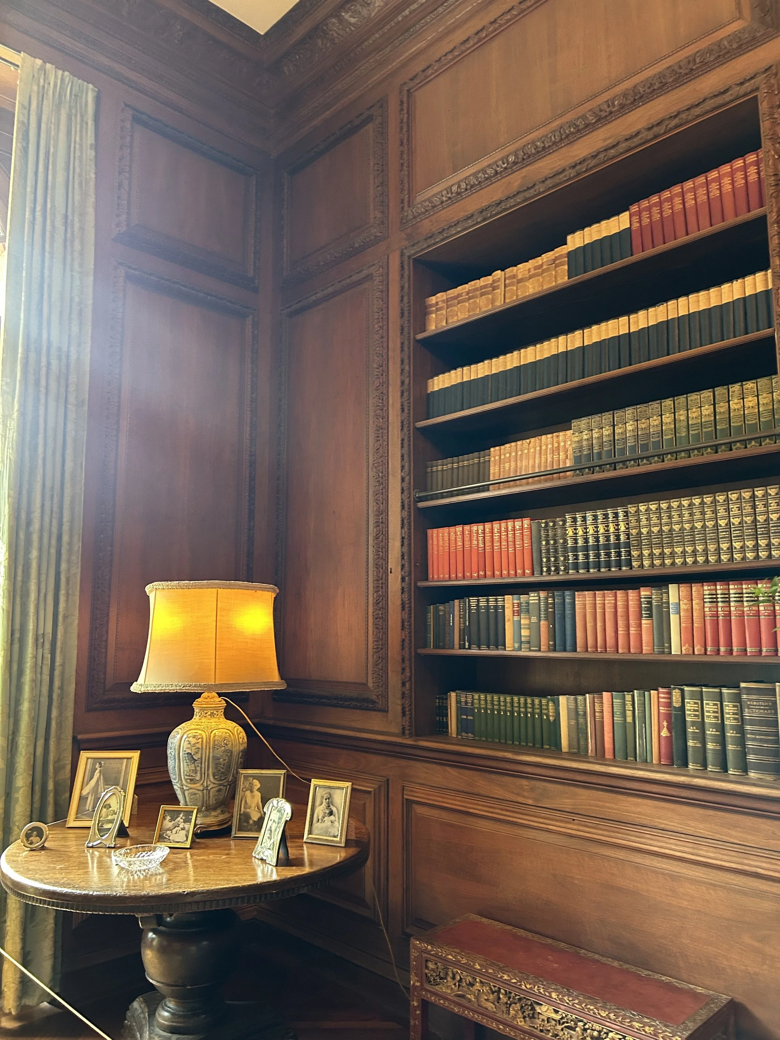 A room with wooden paneled walls and a bookshelf filled with books. There's a side table with framed photographs, a large ornate vase, a lamp with a yellow shade, and a small decorative chest with gold accents.