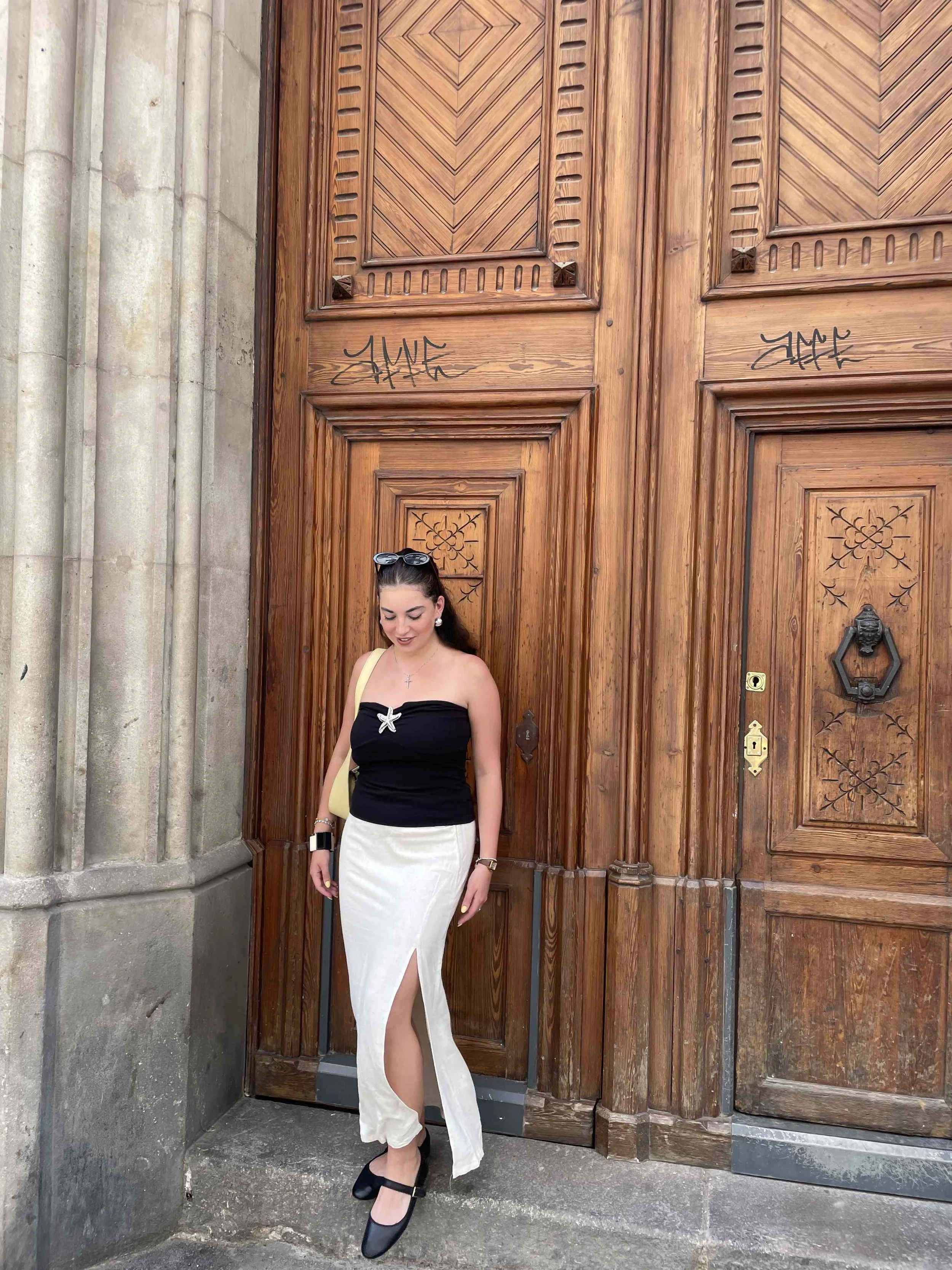 Woman standing in front of a wooden door with intricate carvings, wearing a black strapless top, a white skirt with a front slit, and black shoes. She has sunglasses on her head and jewelry, and is smiling with closed eyes.