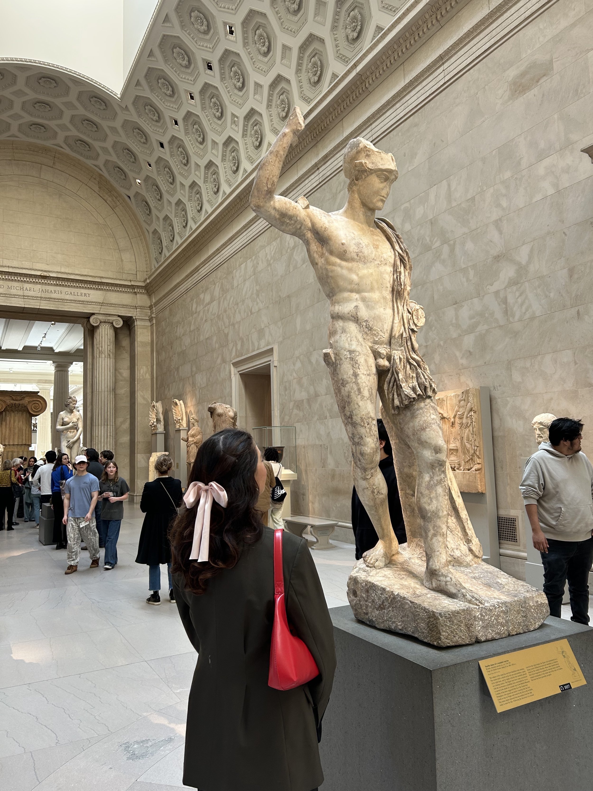 A marble statue of a muscular male figure with a helmet, standing on a pedestal inside a museum, surrounded by visitors. The statue is in a classical style, with a partially damaged arm raised and a cloth draped around his waist. The museum has high ceilings with decorative detailing and walls with informational plaques.