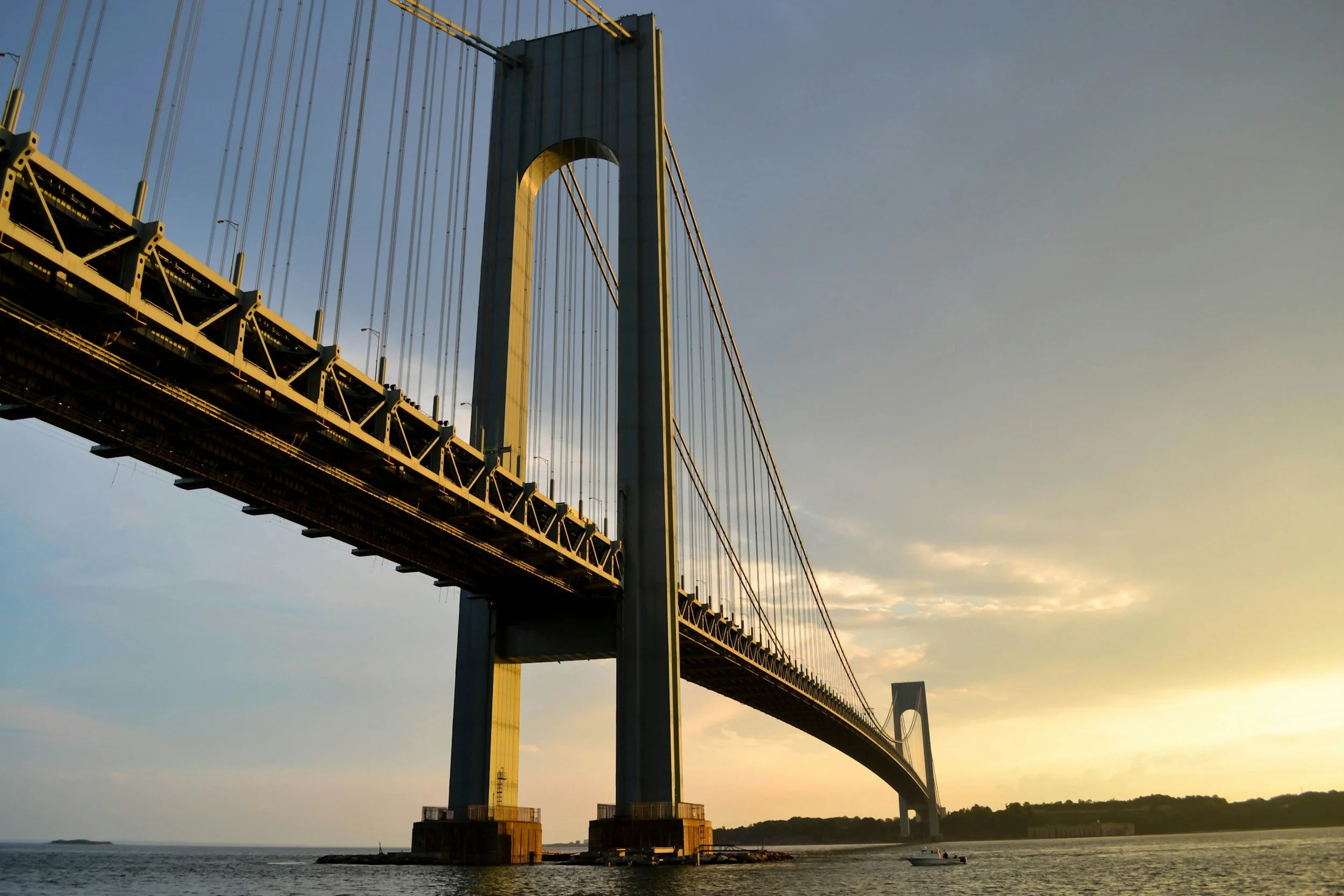 A large suspension bridge spans over water during sunset, with a boat sailing underneath and a distant shoreline in the background.