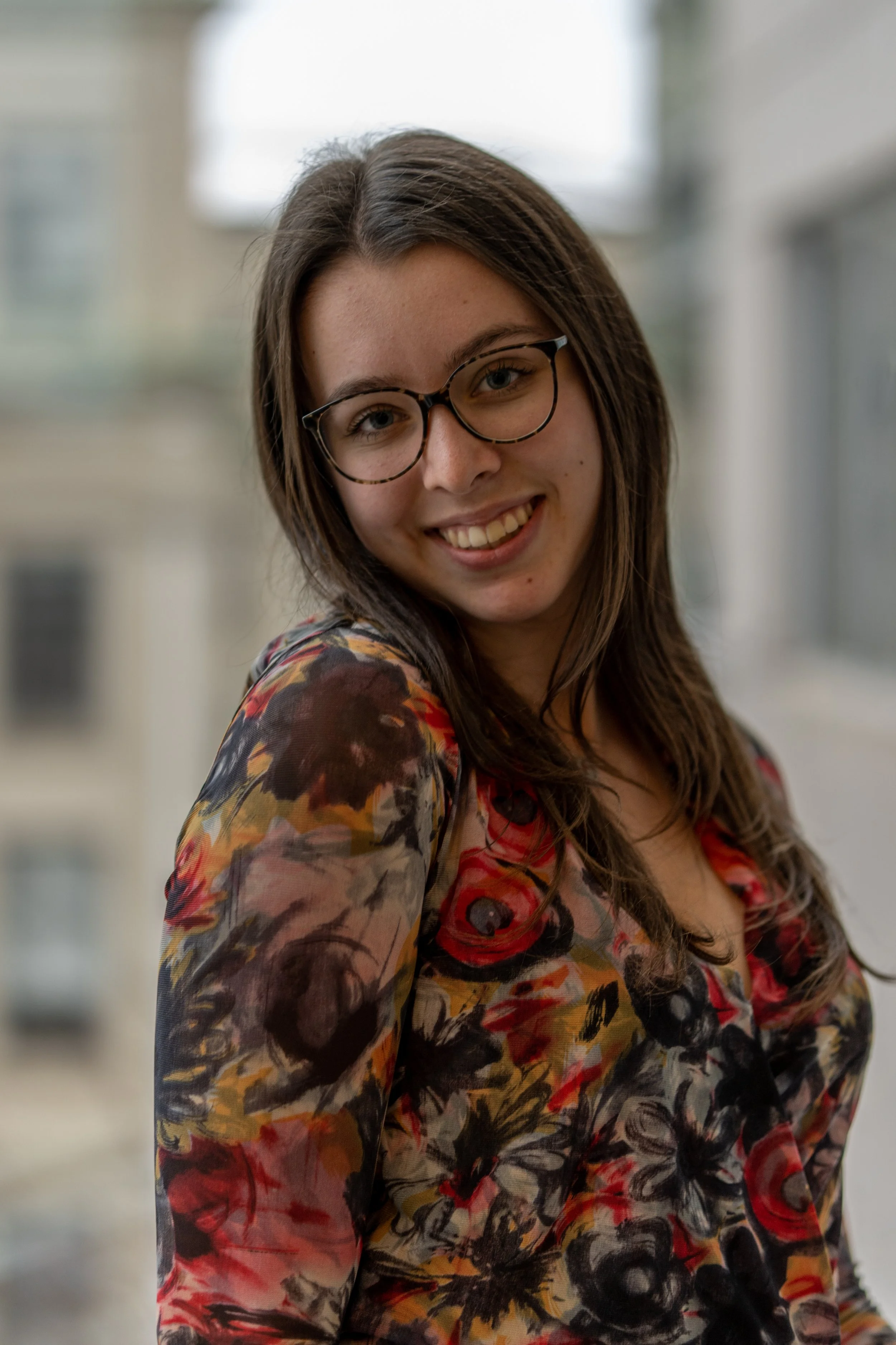 A young woman with long brown hair, glasses, and a bright smile, wearing a colorful floral blouse, standing near a window in an indoor setting.