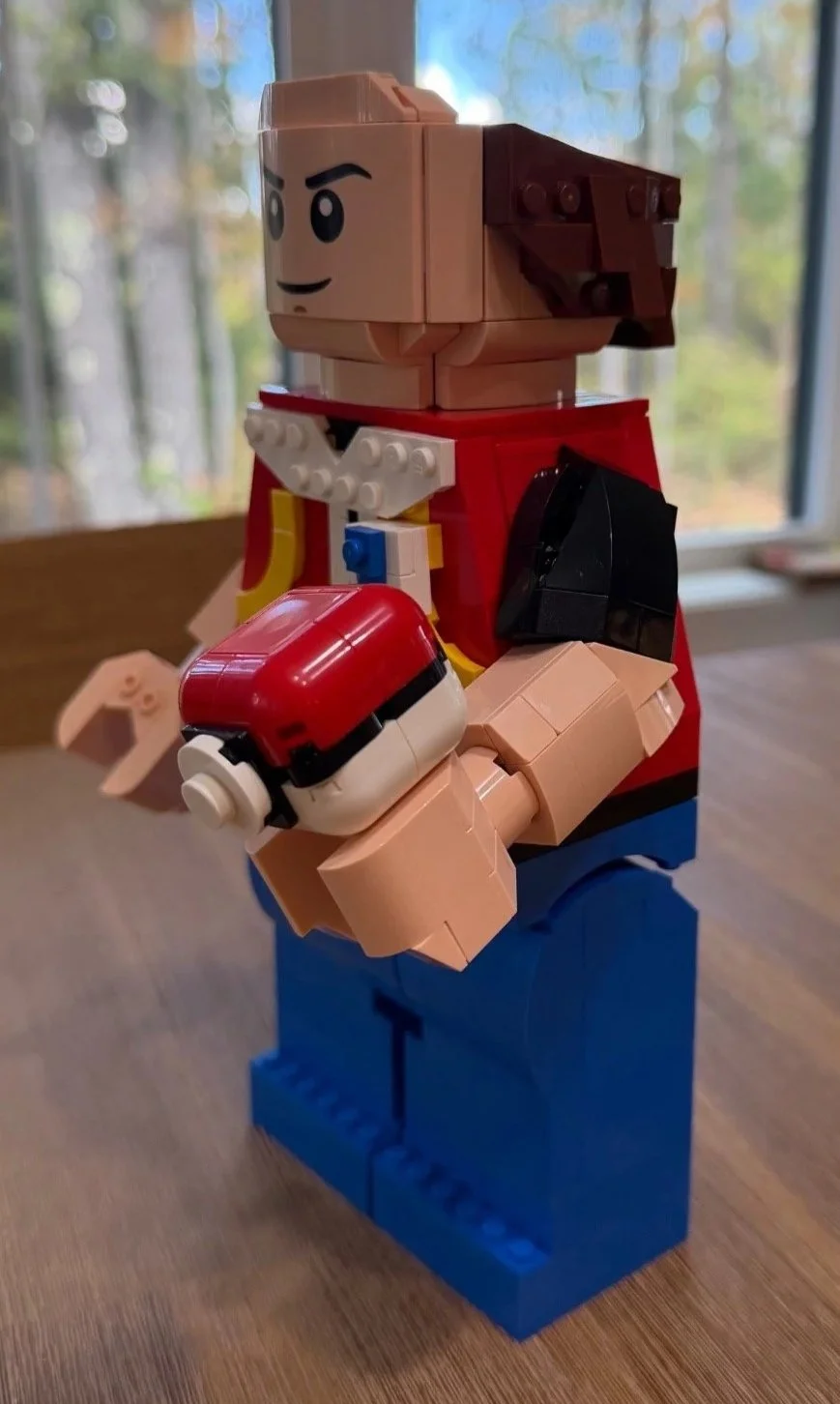 Three-quarter view of LEGO Red holding a red-and-white Poké Ball, with brown hair attached and the cap still absent.