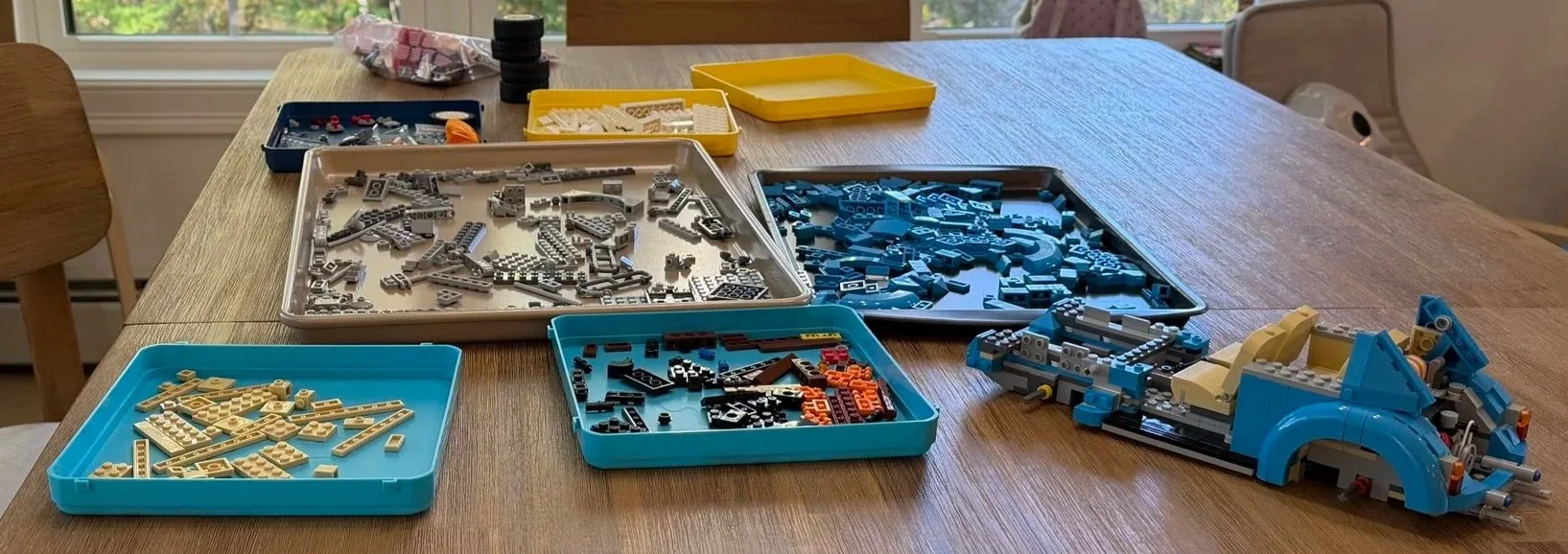 Partly assembled blue LEGO Volkswagen Beetle beside trays of sorted pieces during rebuilding.