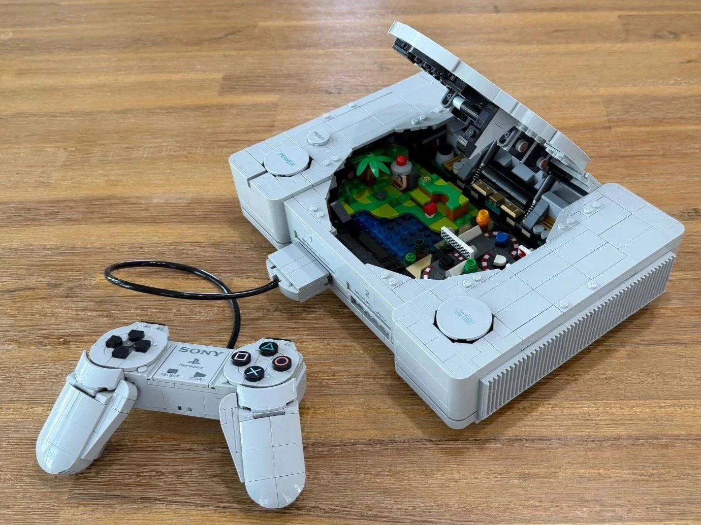 Completed LEGO PlayStation with its lid raised above the two game scenes and its controller connected beside the console.