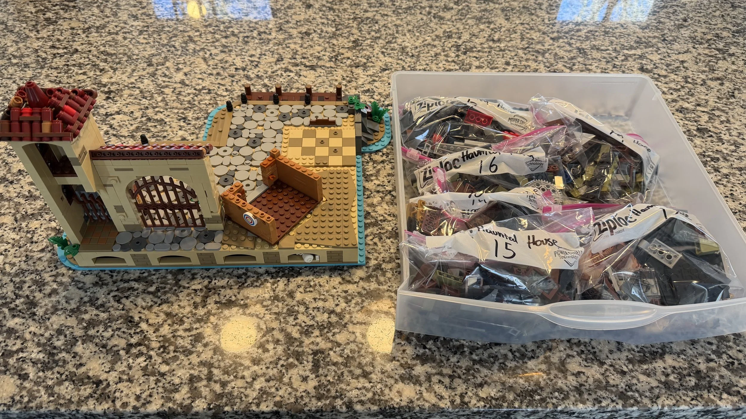 LEGO haunted house under construction on a granite countertop, with a container of LEGO pieces labeled with house numbers.
