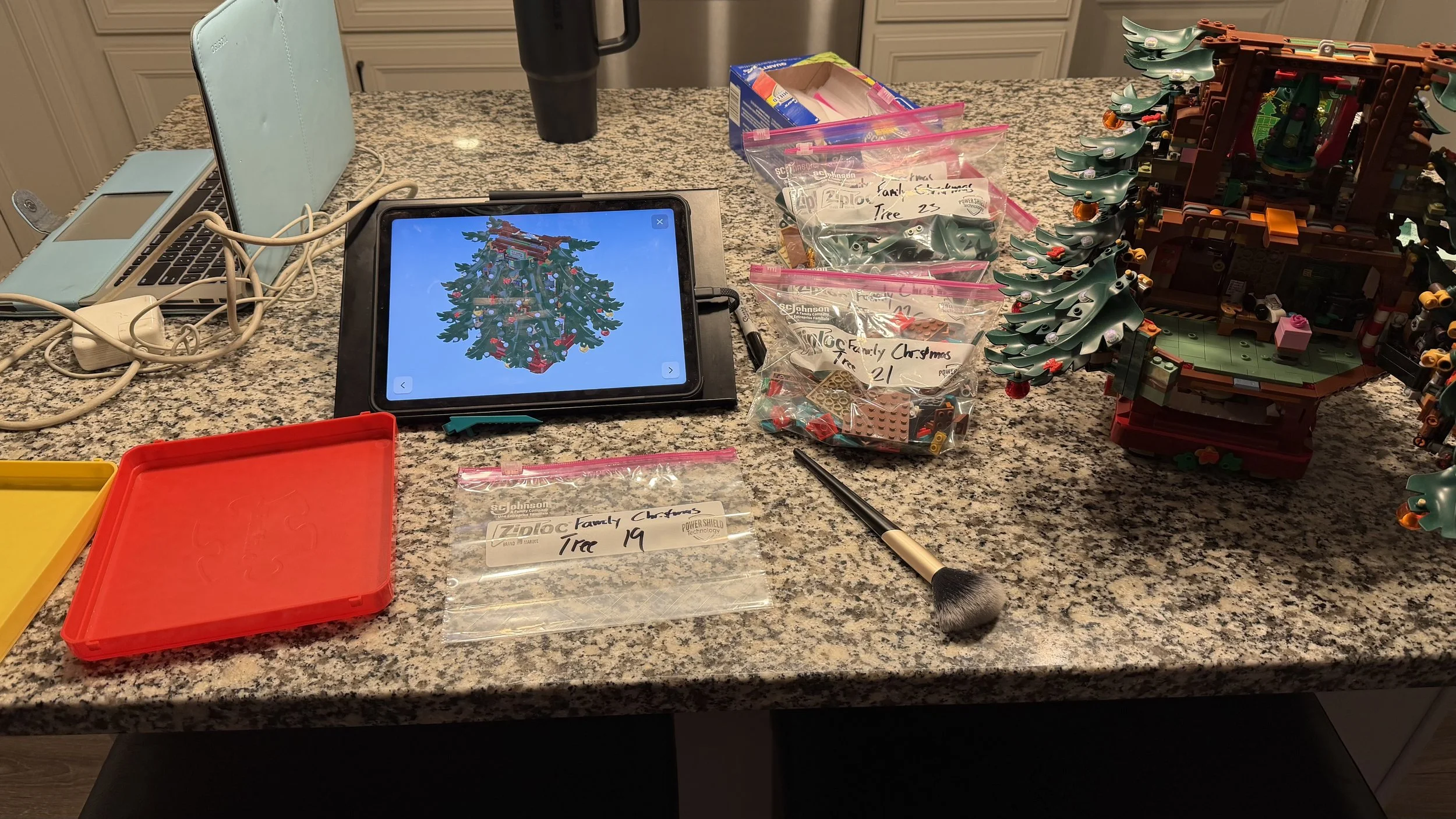 A countertop with a LEGO Christmas tree and a partially assembled LEGO house, plastic bags with LEGO pieces, a tablet displaying a LEGO Christmas tree, a makeup brush, a laptop, a digital clock, and various charging cables.