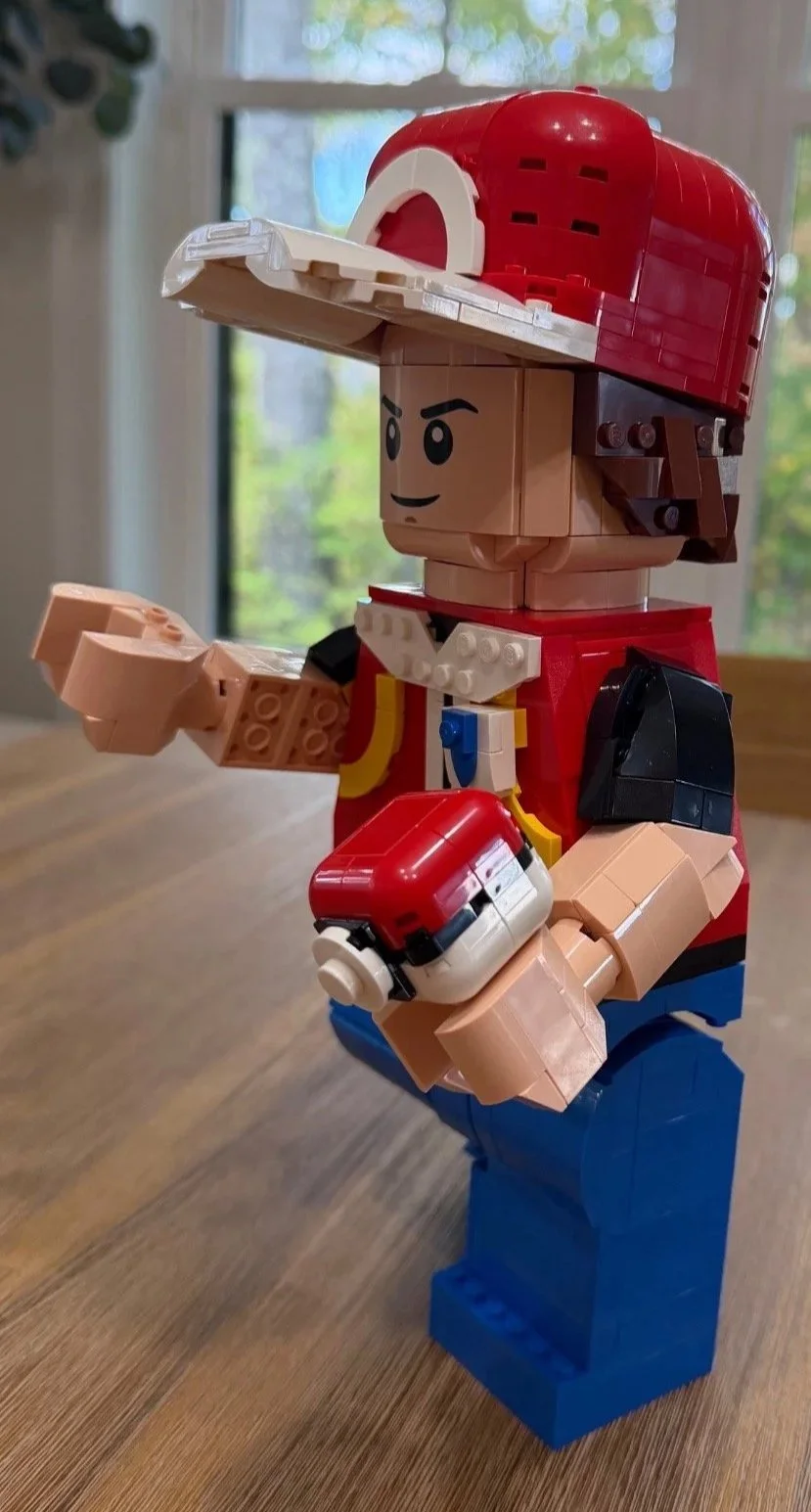 Completed LEGO Red in a three-quarter pose, showing his face, cap brim, black sleeve, large hand, and red-and-white Poké Ball.