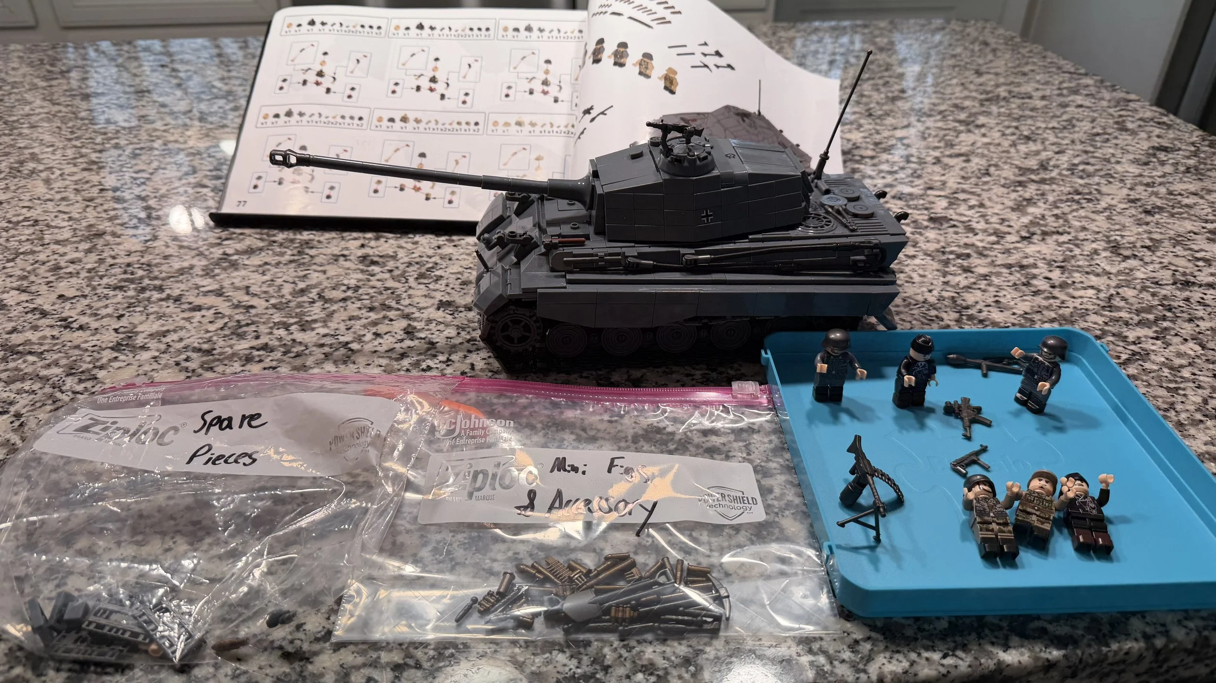 A LEGO tank model displayed on a speckled granite countertop with LEGO minifigures and accessories on a blue tray, an instruction manual in the background, and a plastic bag labeled 'Spare Pieces & Accessories' containing various small LEGO parts.