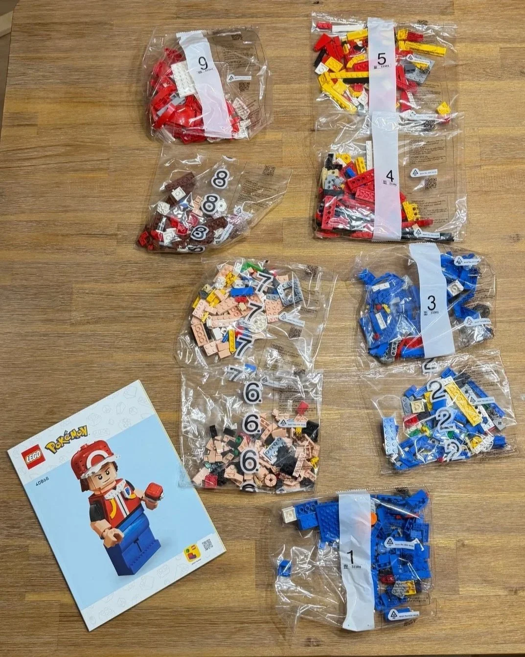 Nine numbered LEGO Up-Scaled Red parts bags and the instruction booklet arranged on a wooden table before building.