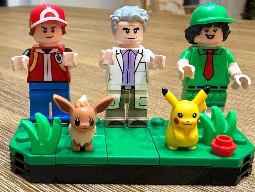 Red, Professor Oak and Picnicker minifigures behind Eevee and Pikachu on a green LEGO display strip.