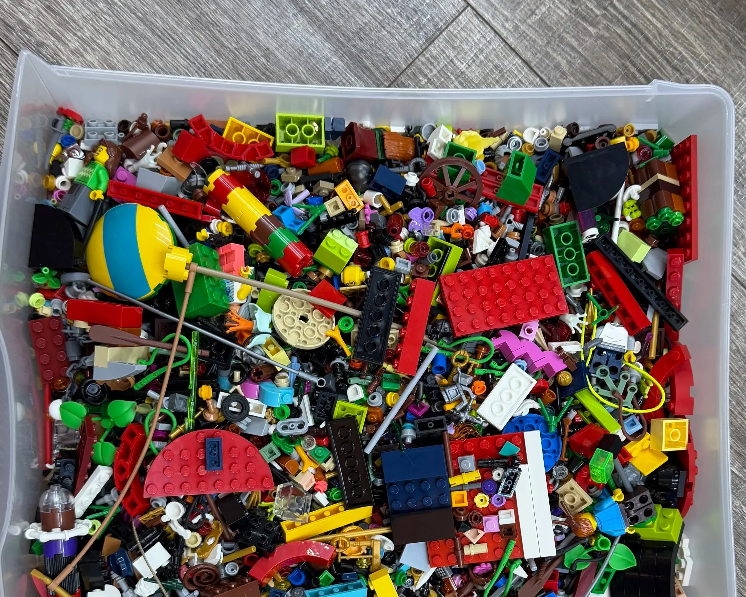 A plastic storage bin filled with assorted LEGO bricks, pieces, and accessories in various colors and shapes.