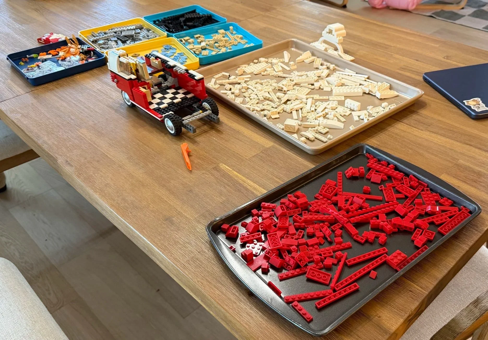 Red LEGO camper van chassis with an exposed checkerboard interior beside trays of red, white and other pieces.