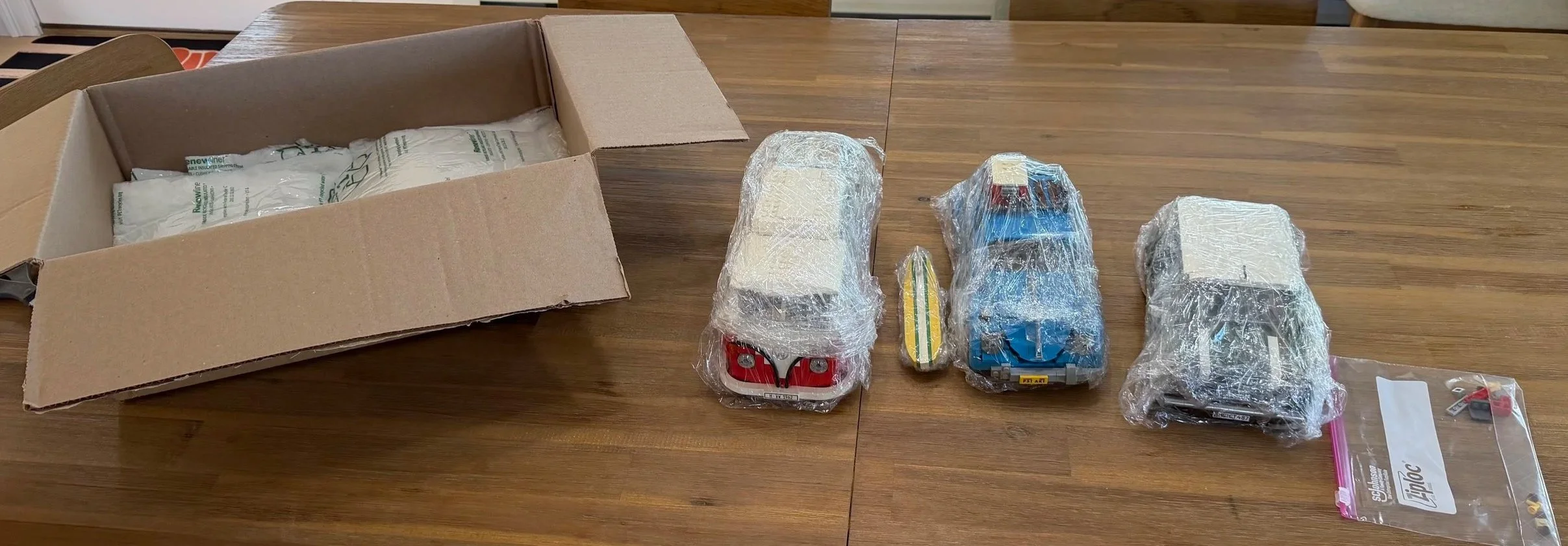 Three rebuilt LEGO vehicles wrapped on a table beside a separate surfboard, a bag of extra pieces and an open box.