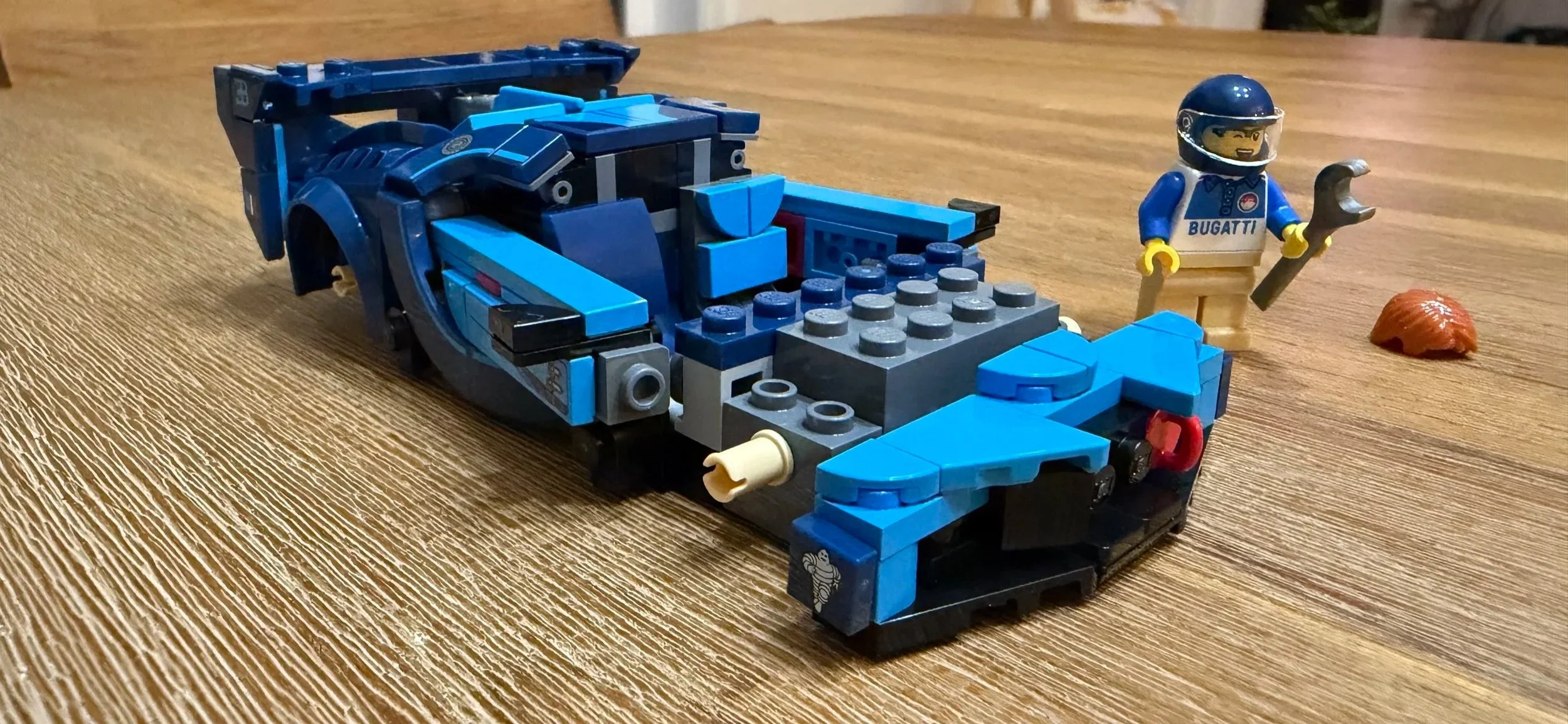 Unfinished LEGO Bugatti with a blue front bumper, printed Michelin Man detail, exposed hood structure, and driver alongside.
