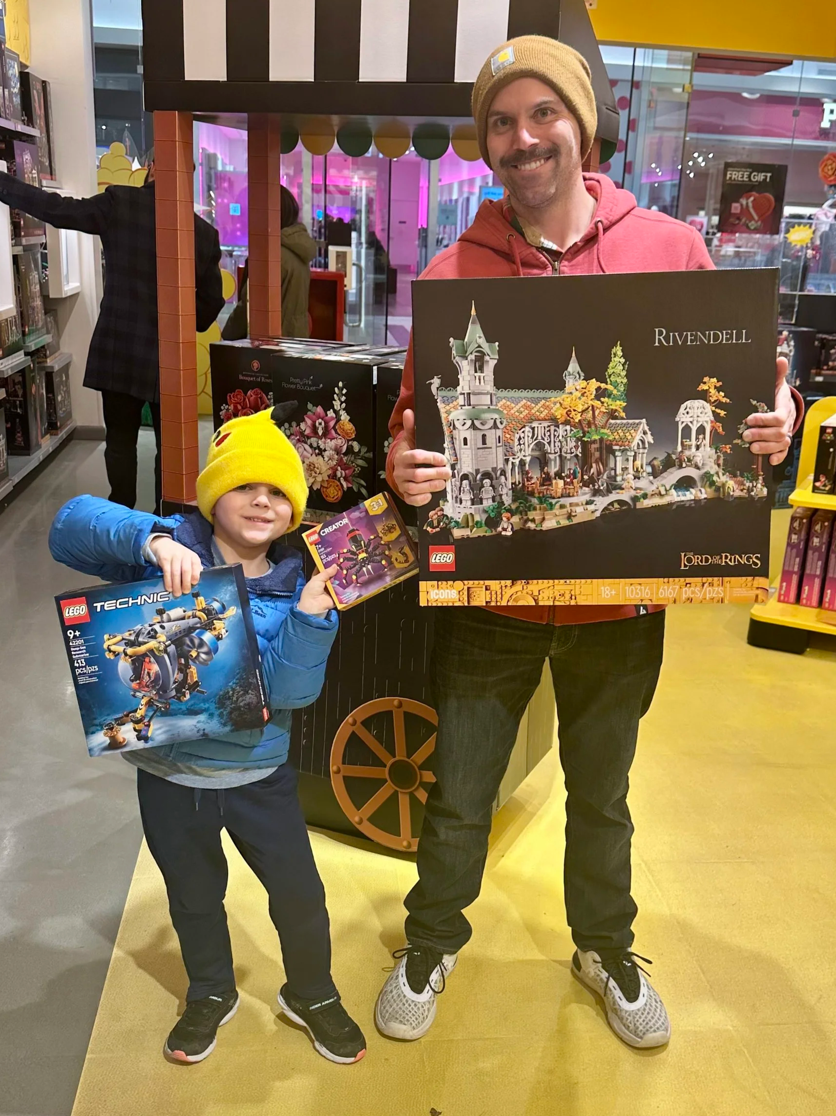 A man and a young boy standing inside a store, holding Lego sets and a Lord of the Rings playset. The man is smiling and wearing a beige beanie and red hoodie, while the boy is smiling and wearing a yellow beanie and blue jacket.