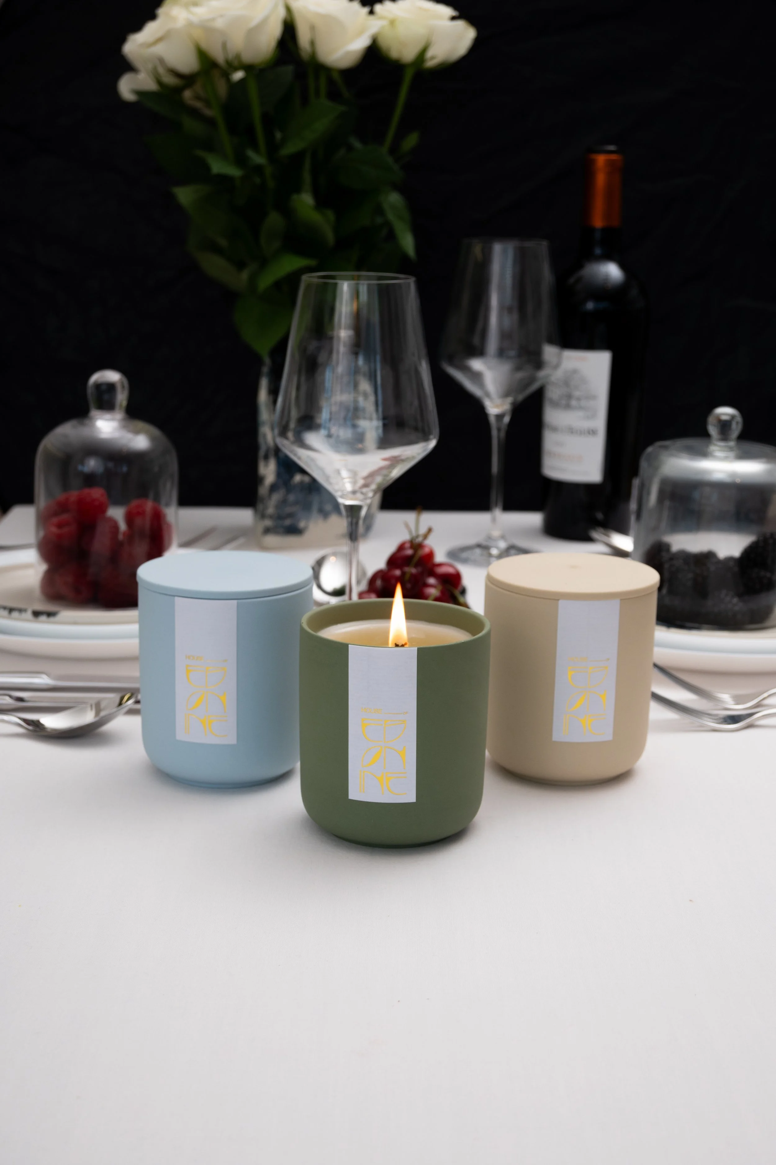 Elegant dining table setting with three colored candles, wine glasses, a bottle of wine, and decorative glass covers over fruit, with a flower and black background.