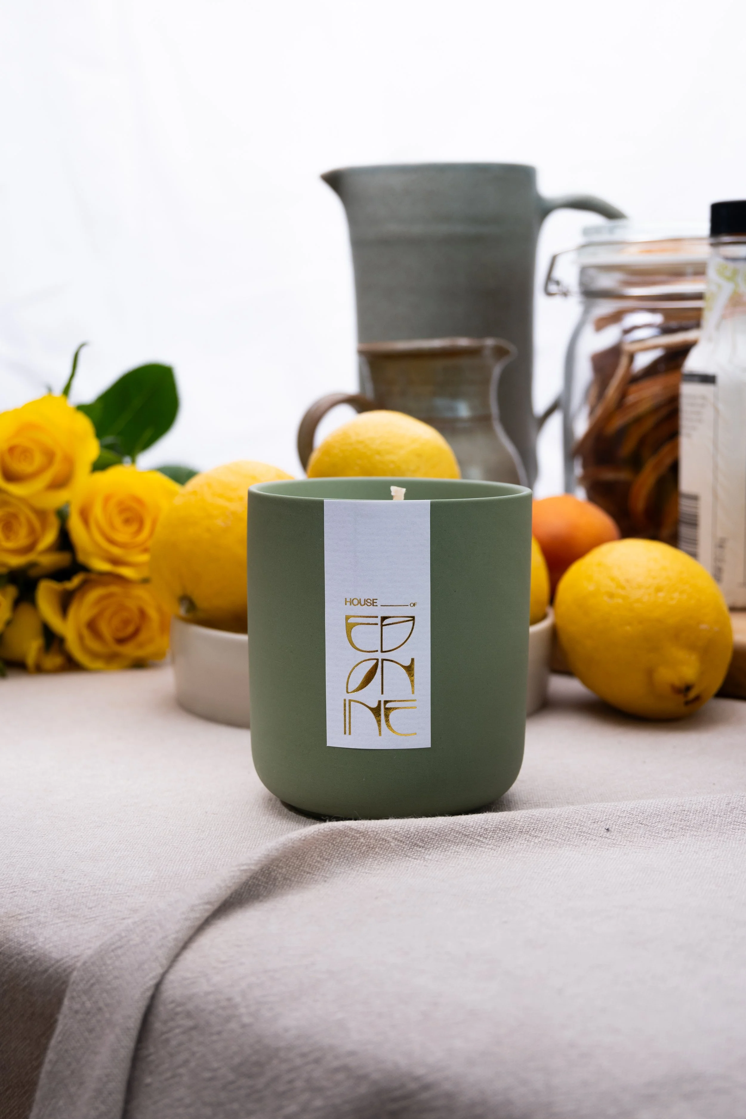 A green candle with a gold and white label that reads 'HOUSE OF GARDEN' placed on a table with lemons, yellow roses, and kitchenware in the background.