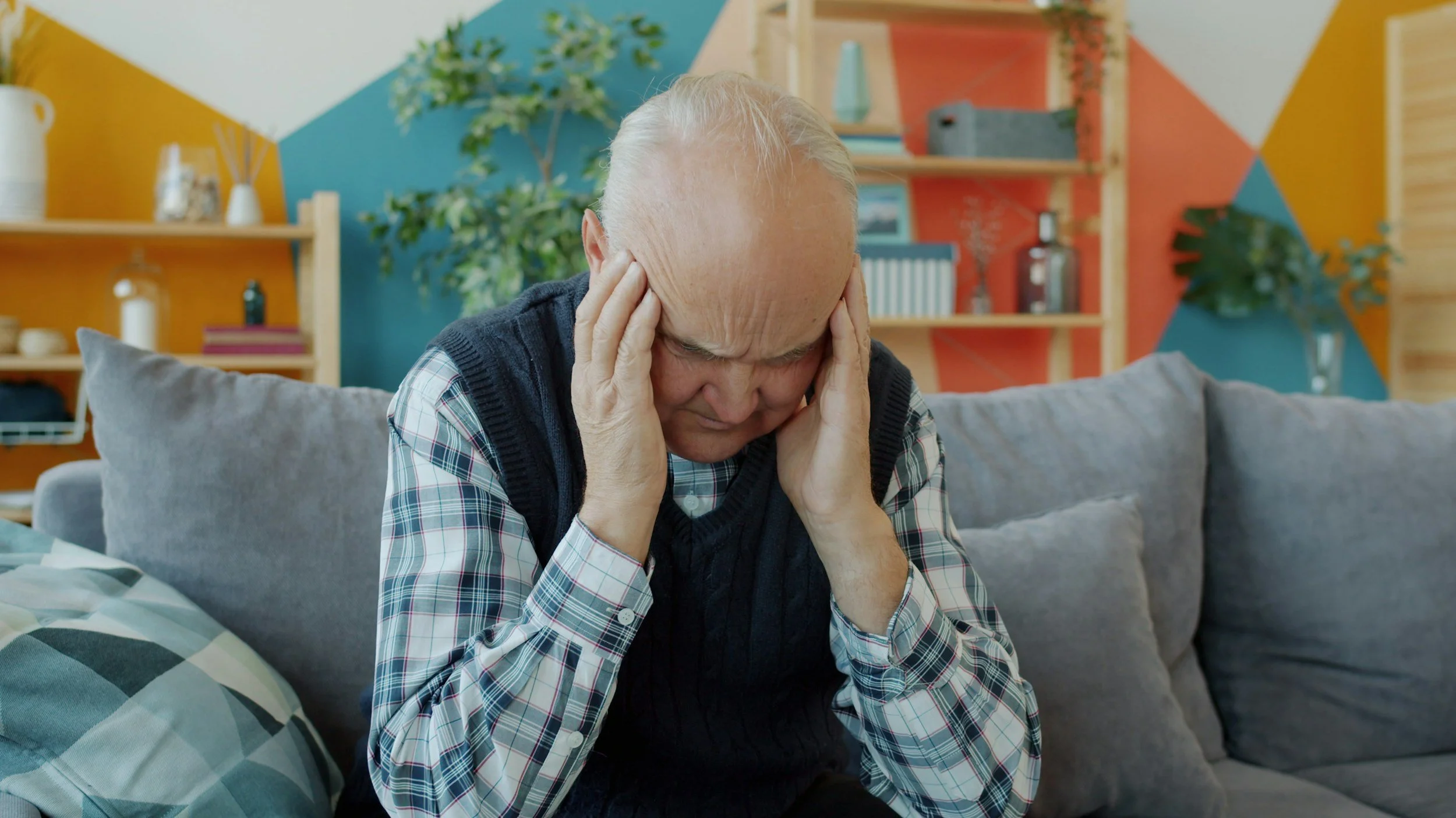 An elderly man sitting on a couch, holding his head with both hands, appearing distressed or in pain, in a colorful living room with decorative shelves and plants in the background.