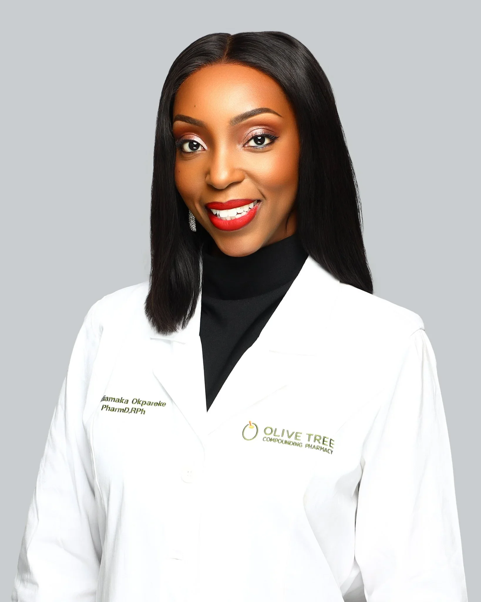 A woman with long black hair, red lipstick, and a black turtleneck, smiling, wearing a white lab coat with green text that reads 'Olive Tree Compounding Pharmacy' and her name and credentials.