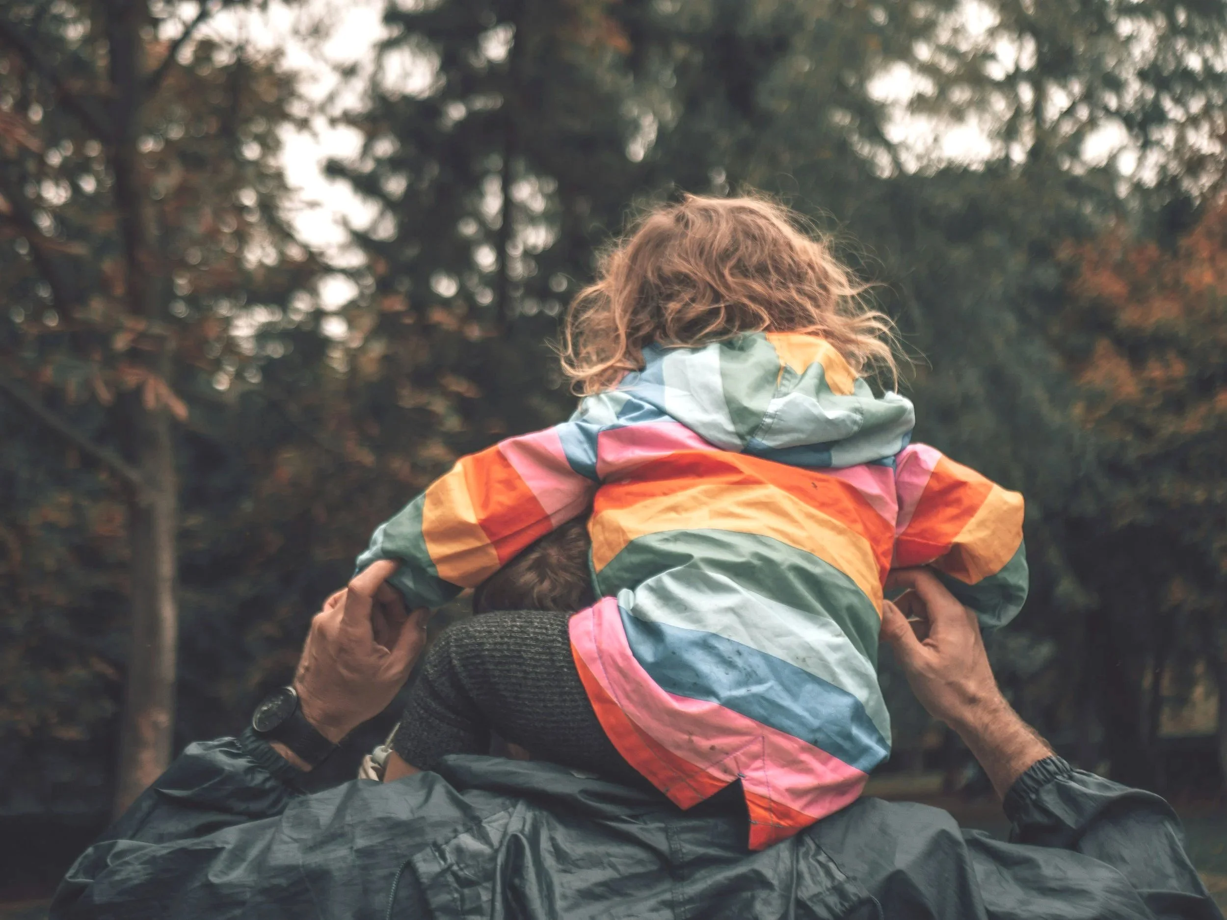 A person with curly hair wearing a rainbow-colored jacket holding a child with dark hair. The child has their head resting on the person's shoulder. They are outdoors with trees in the background.
