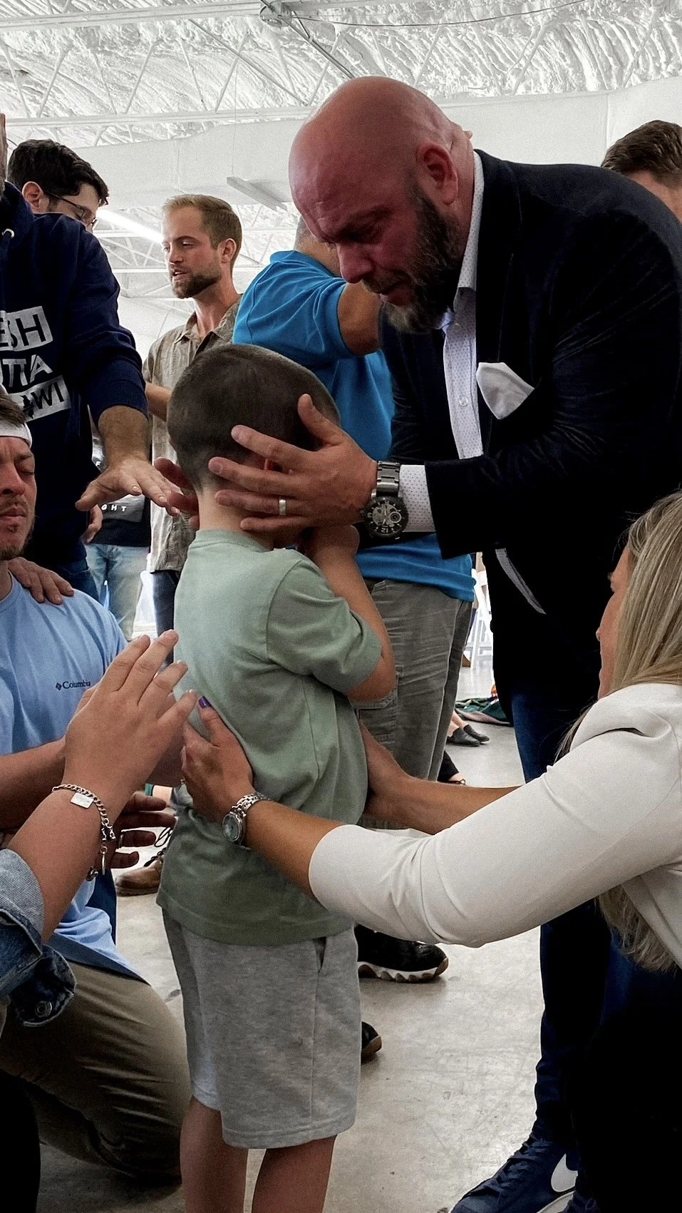 A group of people gathered around a young boy, who is standing with his eyes closed, while an older man in a suit and a woman in white help him pray or have a moment of reflection. The setting appears to be indoors, possibly at a community or religio