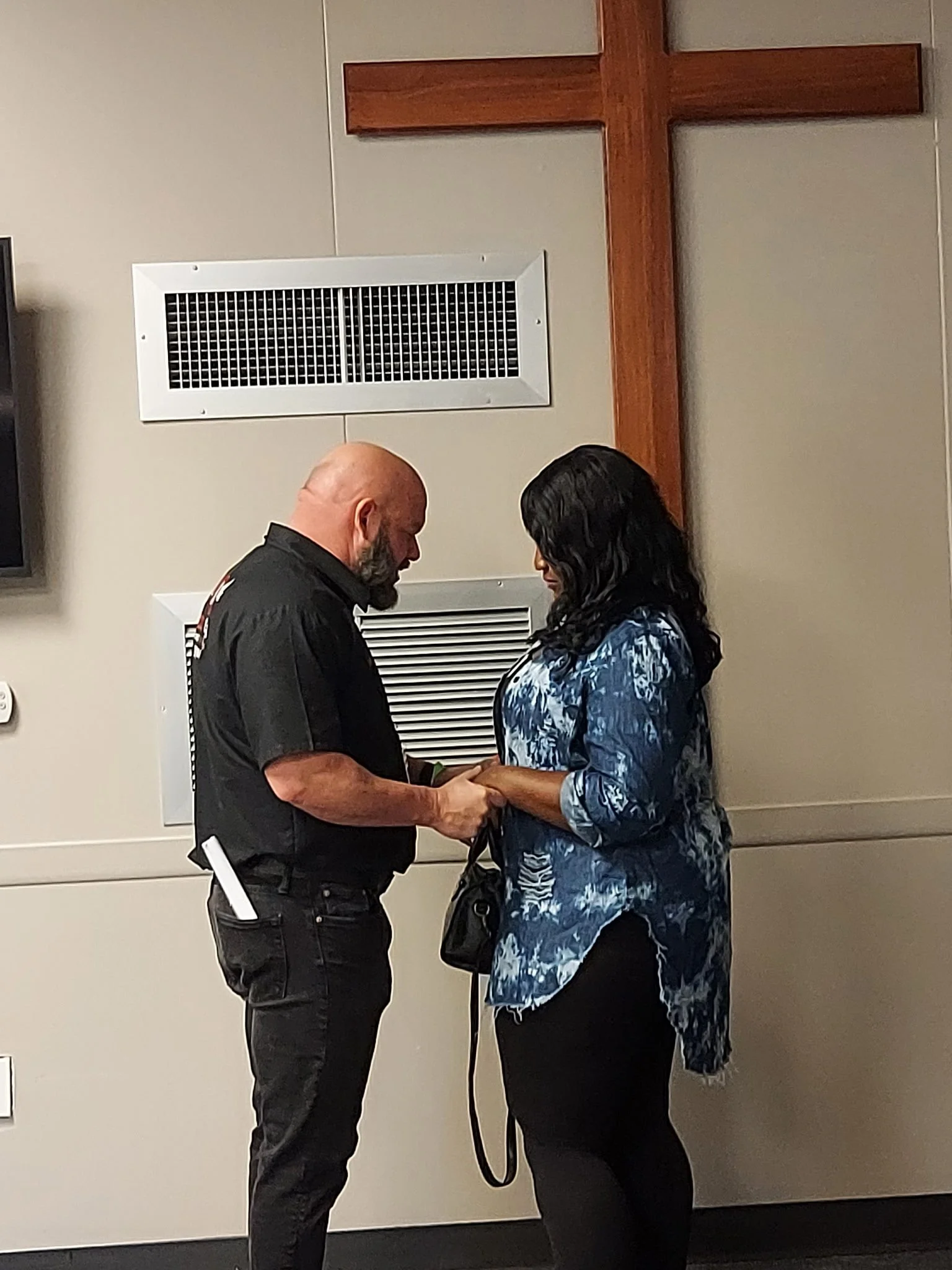 A man and woman holding hands and looking at each other inside a room with a large wooden cross on the wall.