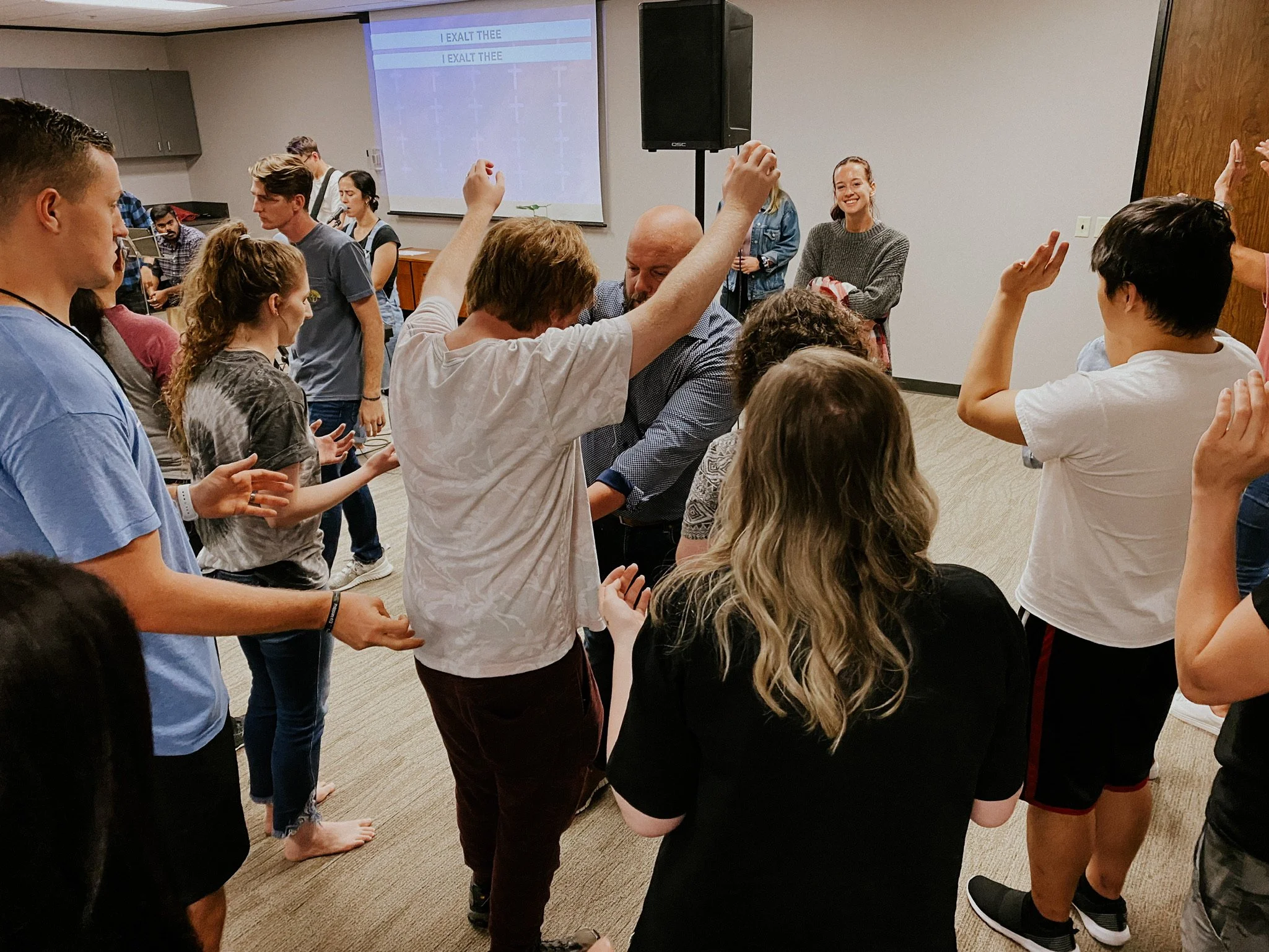 A group of people standing in a circle in a room, some are raising their hands, with a few smiling and engaging in a group activity or discussion.