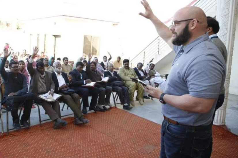 A man with glasses and a beard wearing a gray polo shirt, standing in front of an audience, raising his right hand while holding a tablet in his left hand. The audience, mostly men in formal attire, are seated and raising their hands in response, during a presentation or seminar in an indoor setting with white walls and stairs in the background.