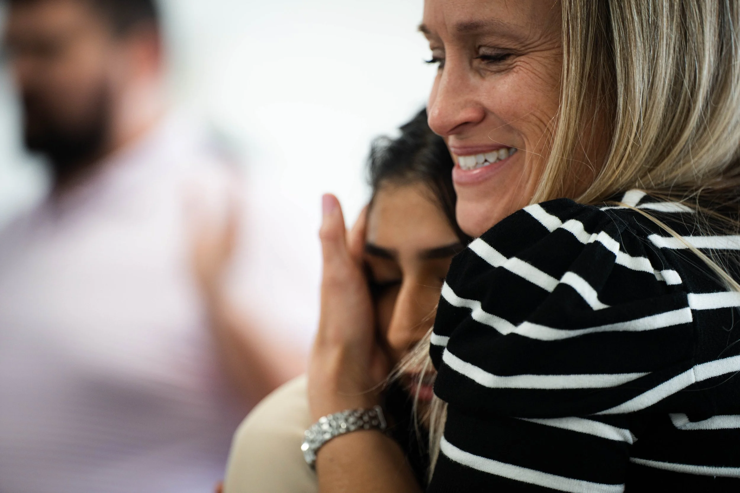 A woman smiling embraces a young girl with her eyes closed, in a close-up indoor setting with two blurred people in the background.