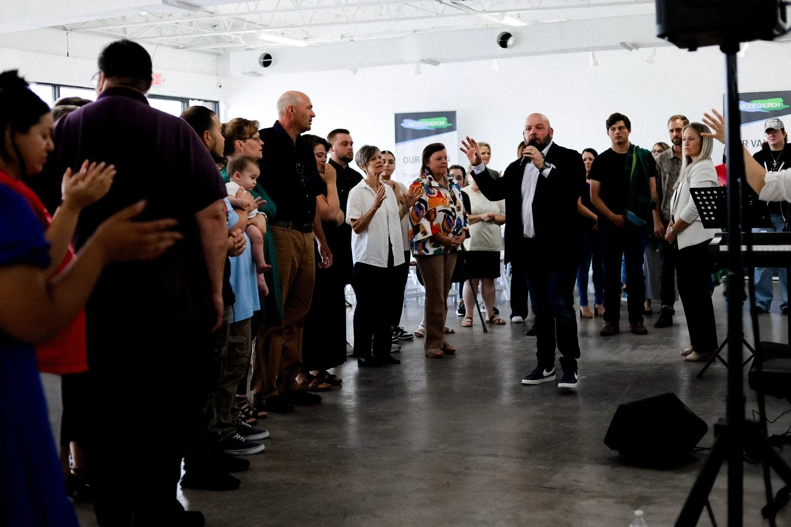 A diverse group of people standing in a circle inside a modern, well-lit room, with one man speaking into a microphone, likely at a church or community event.