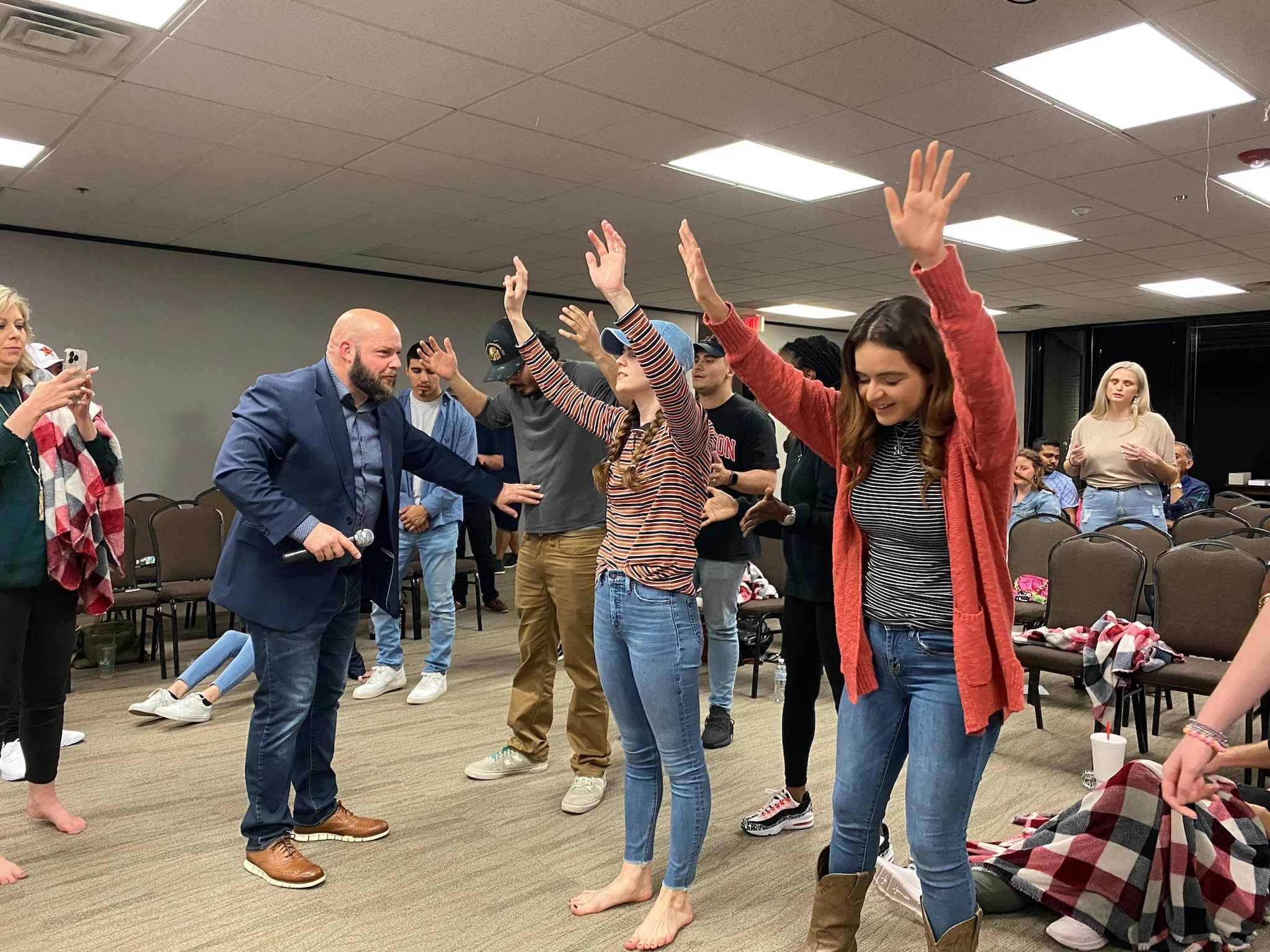 Group of people participating in a prayer or spiritual activity with hands raised, in a room with chairs and casual attire.