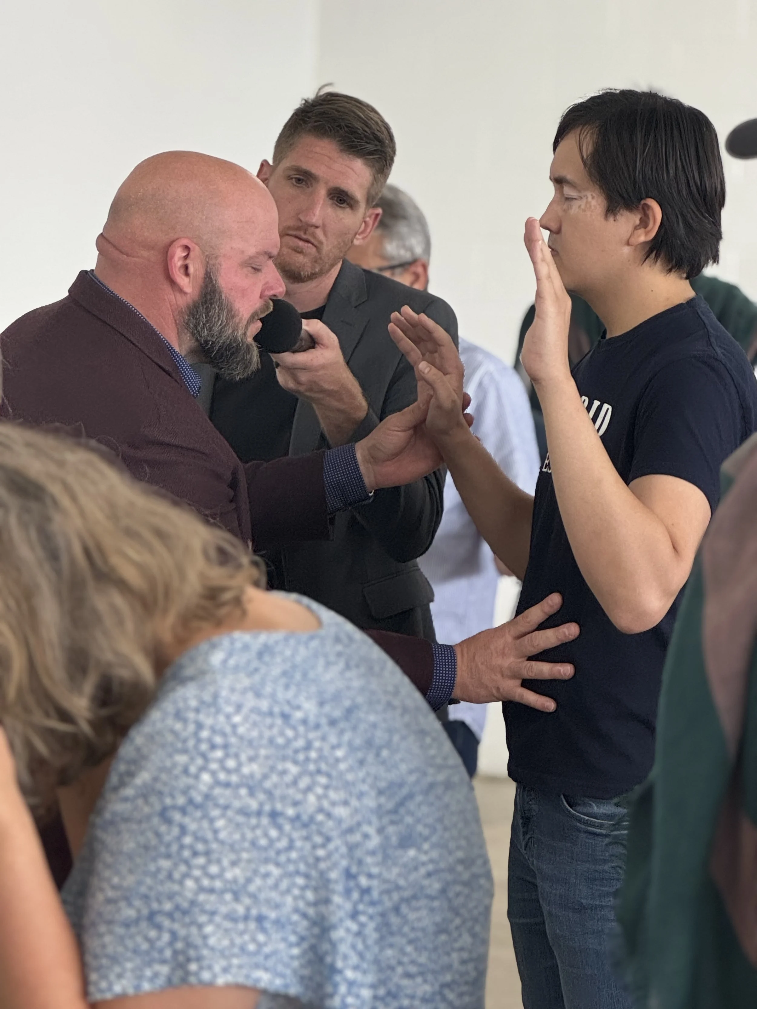 A man with a beard and a maroon jacket is praying with a young man in a navy T-shirt, with his hands raised and eyes closed. The young man appears to be receiving a blessing or prayer from the older man. There are other people around them in a group 