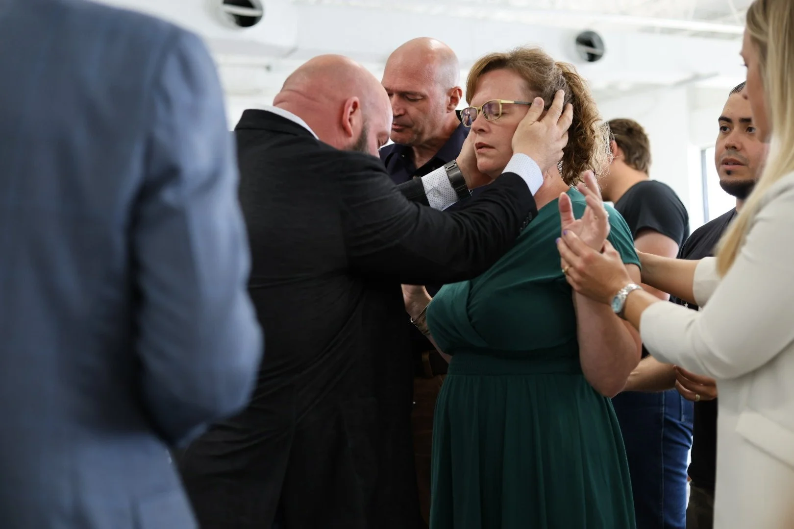 A group of people participate in a religious handshake ceremony at a church. A man is performing the blessing over a woman in a teal dress as others look on.